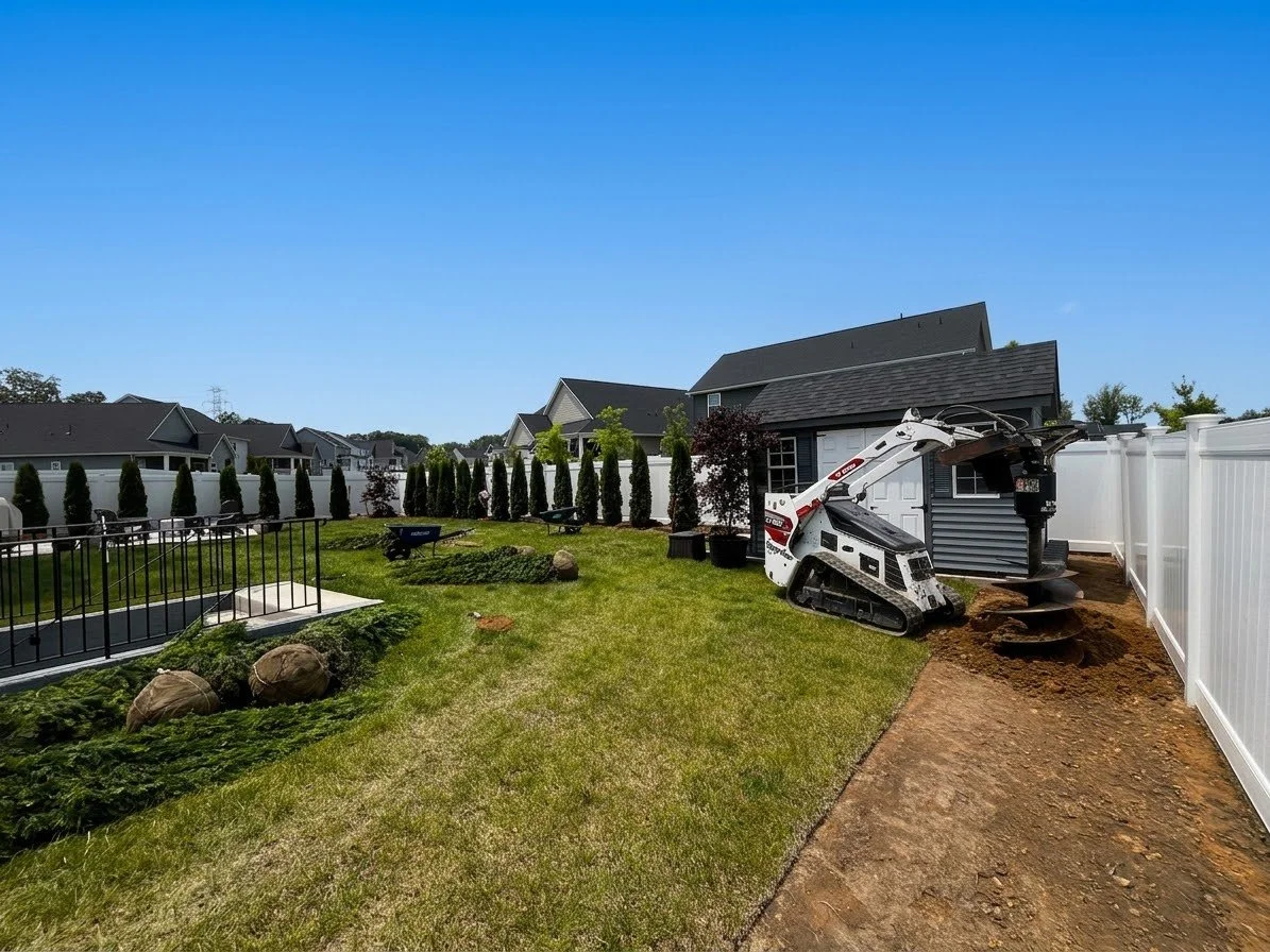 Backyard with a white and black mini excavator digging near the house, with a white fence, grassy lawn, young trees, and neighboring houses in the background.