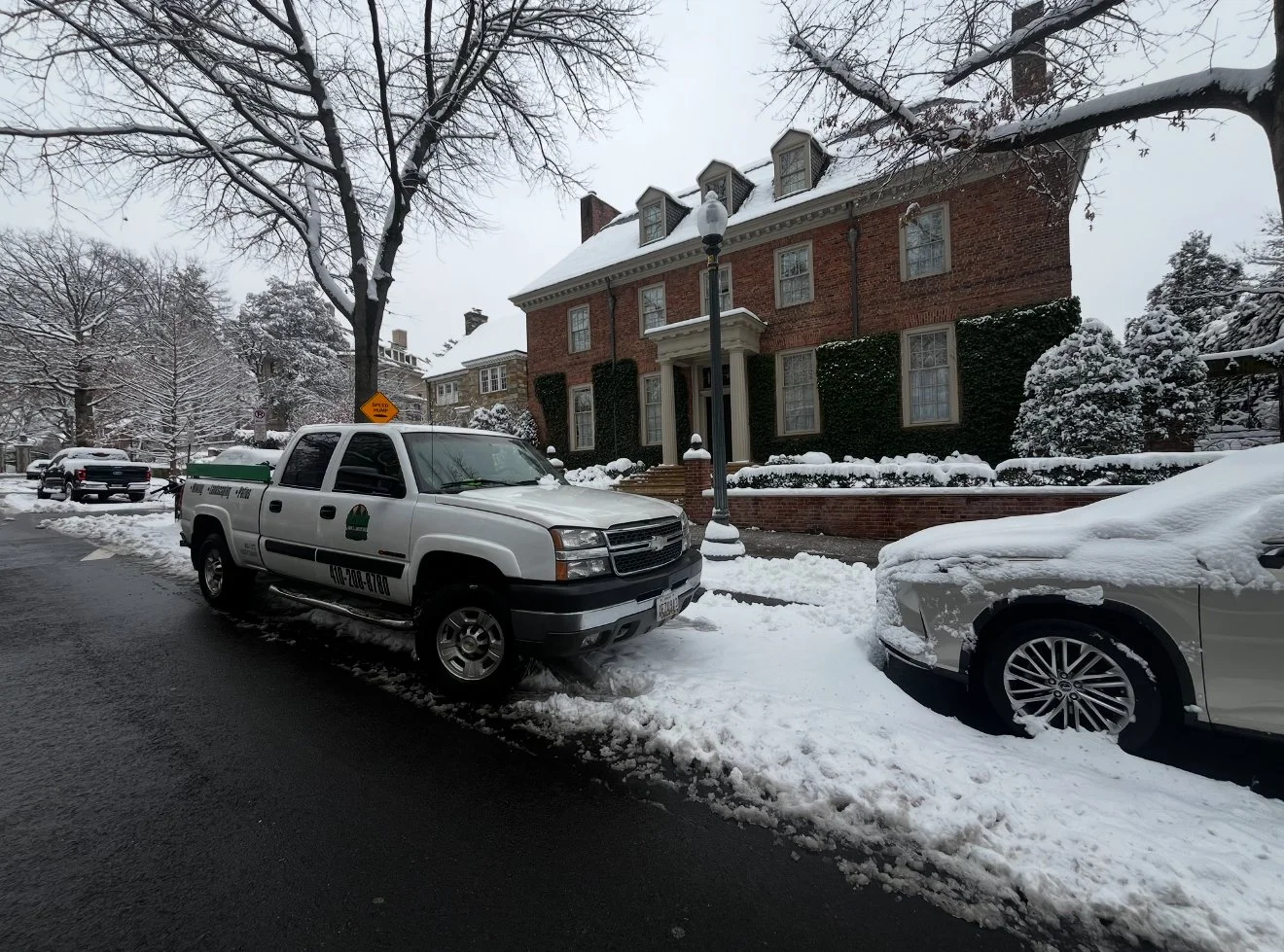 Snow-covered street with parked cars, including a white pickup truck with a green logo and a white sedan with snow on the hood and wheel, in front of a large brick house with ivy-covered walls and a snow-covered yard, during winter day.