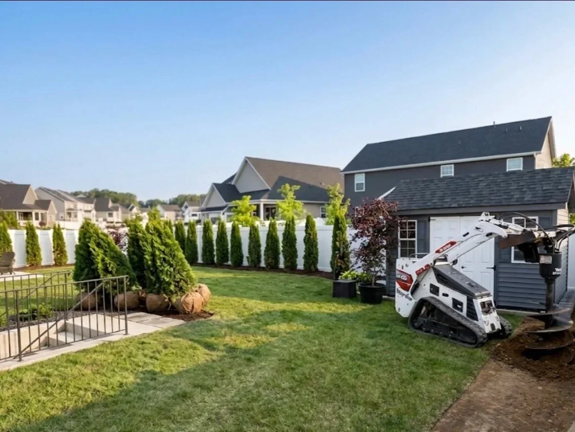 A backyard with green grass, small trees, a white privacy fence, and a small construction excavator near the house.