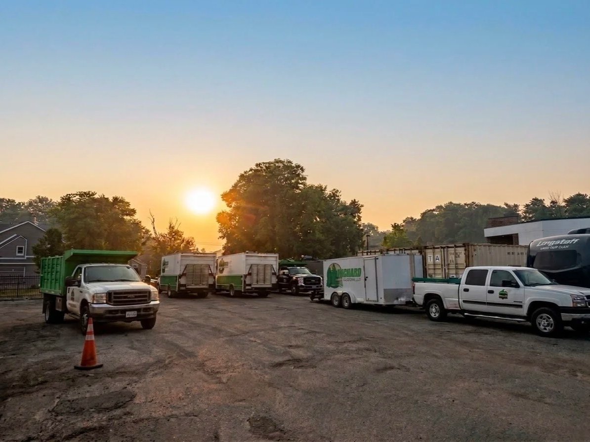 Construction vehicles and trucks parked on a dirt lot at sunrise or sunset, with trees and buildings in the background.