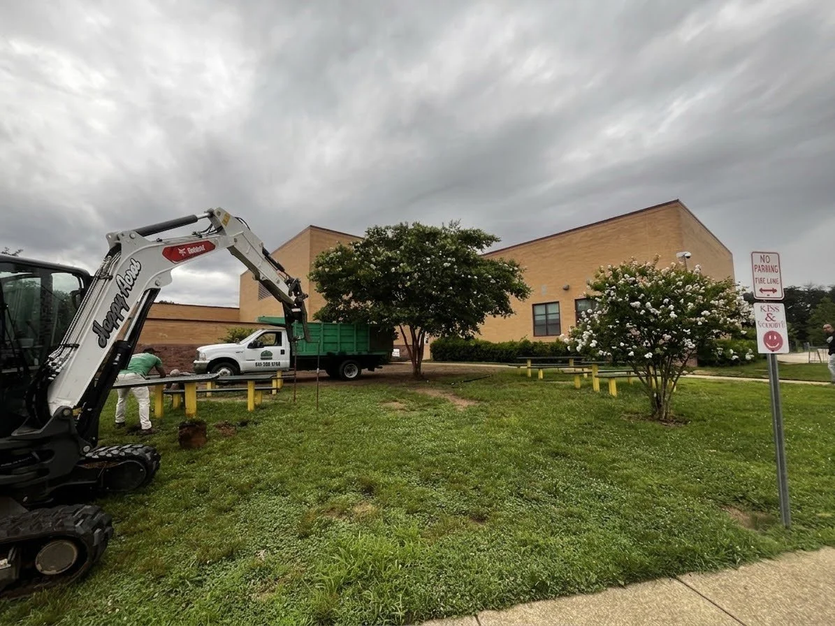 A small green excavator with a white arm near a grassy area, two trees with white flowers, a brown building, a white pickup truck, and a parking sign that reads 'No Parking, Fuse Lane, Kids & School Bus' on a cloudy day.