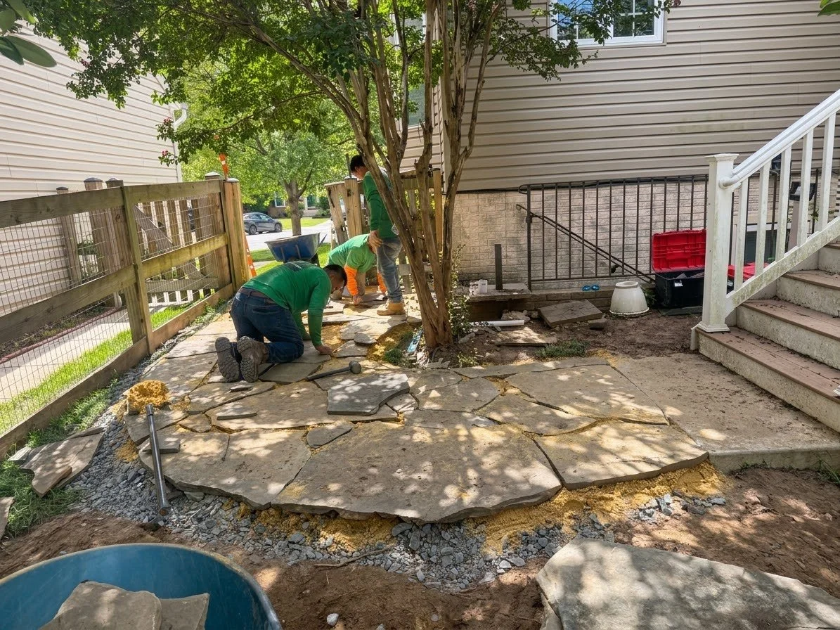 People installing stone pavers in a backyard near a tree, with a house, steps, and fencing in the background.