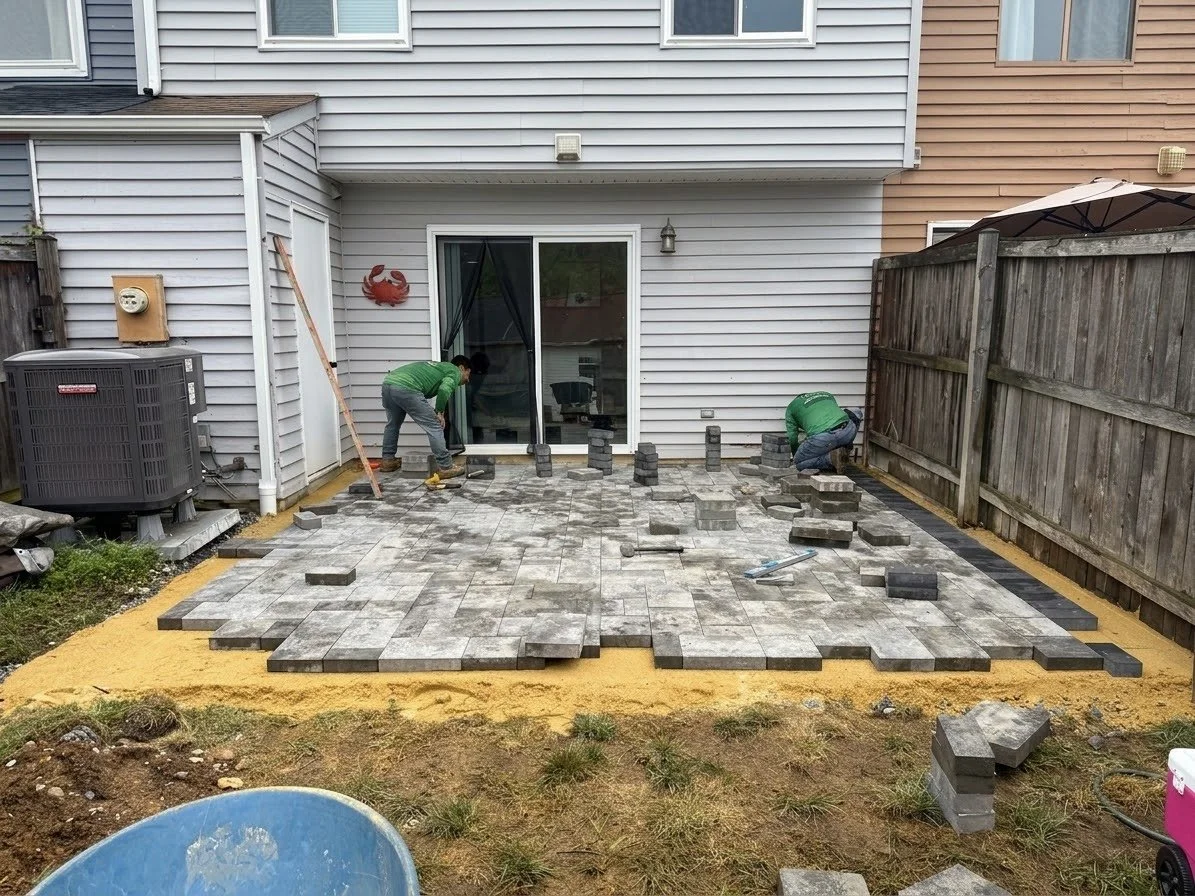 Two workers laying interlocking paving stones on a backyard patio under construction, with some stones stacked nearby, a fence on the right, and a house with a sliding door in the background.
