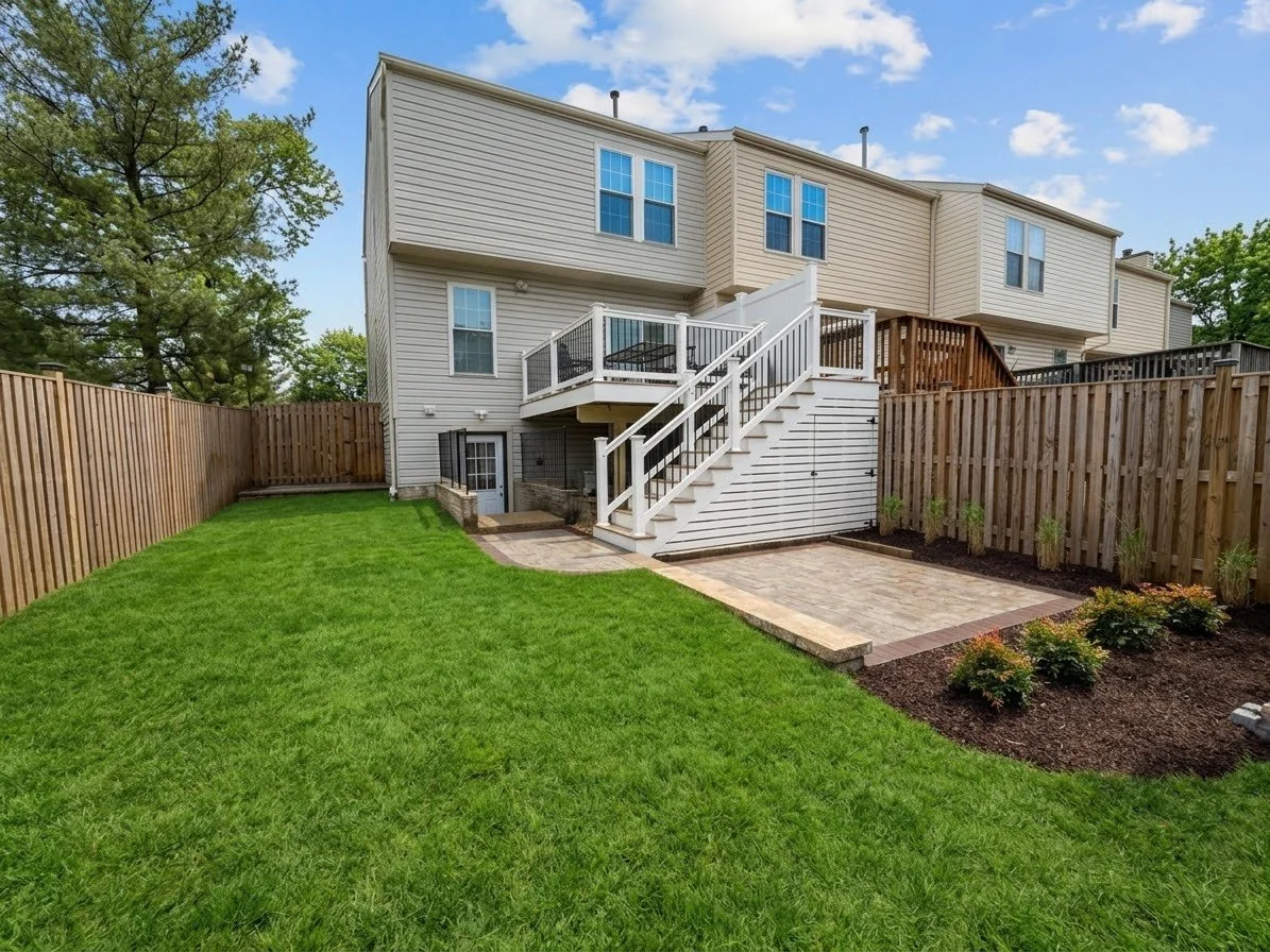 Backyard with green lawn, paved patio area, and wooden fence, with a multi-story house and trees in the background.