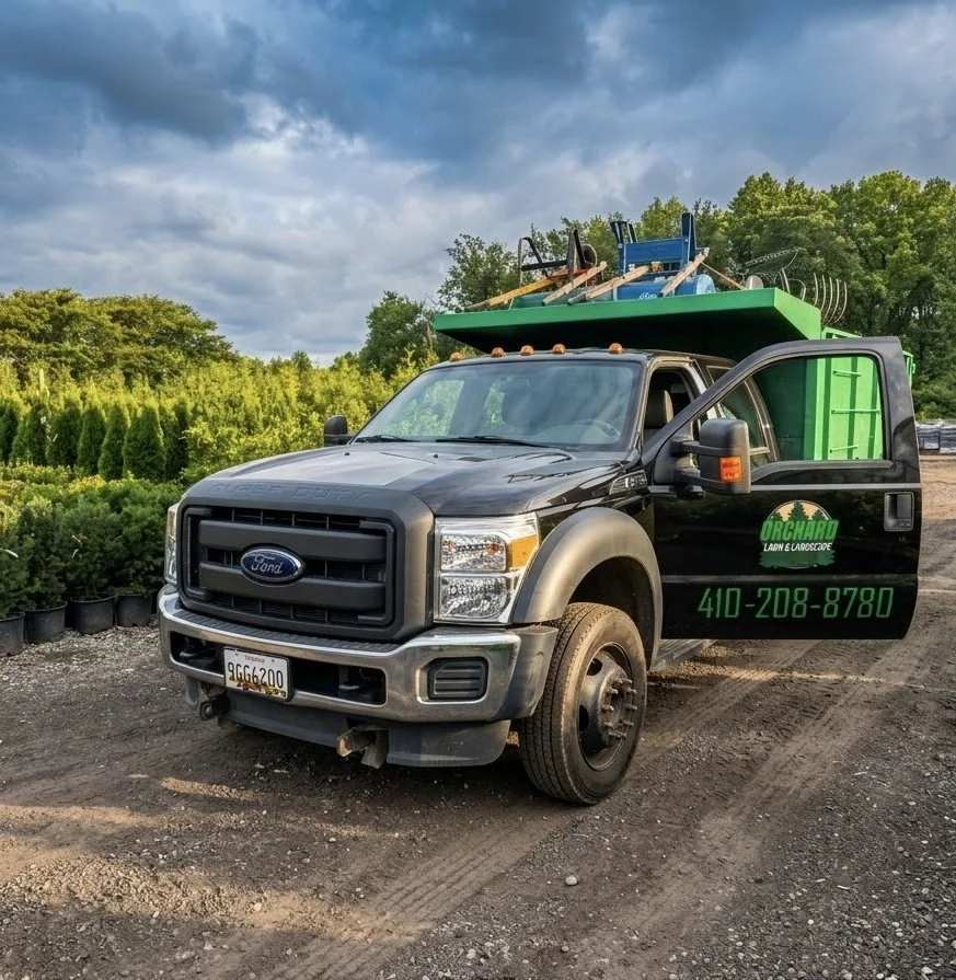 A black Ford truck with a sign for Orchard Lawn & Landscape, including a phone number, parked on a dirt lot near rows of potted plants. The truck is loaded with outdoor equipment on its roof, and the sky is partly cloudy with green trees in the background.