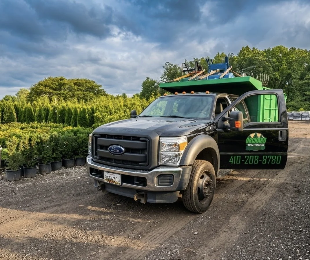 Black Ford truck with a green and black service logo on the door, parked on a dirt path surrounded by potted plants, with a landscape of trees, bushes, and a cloudy sky in the background.