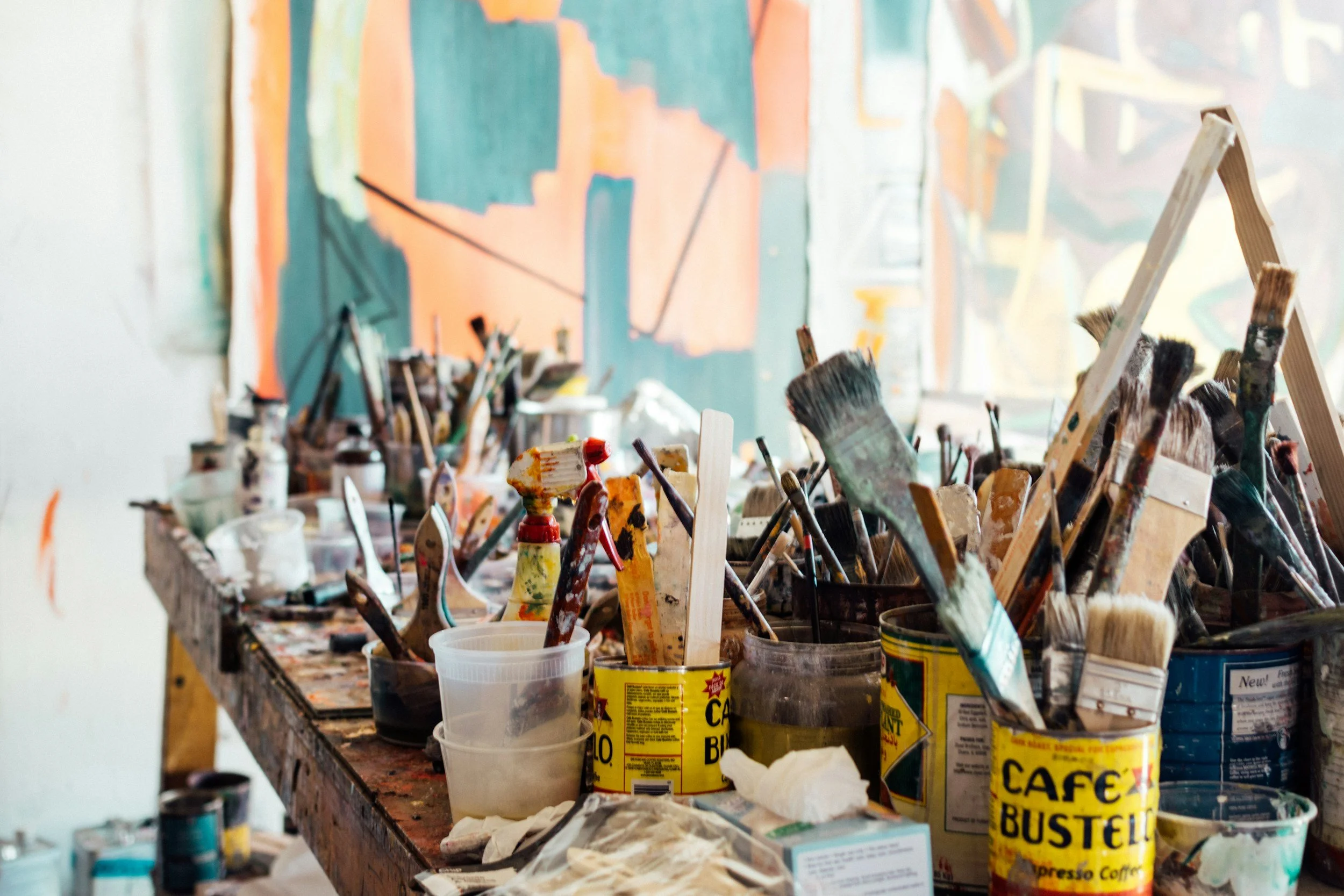 A cluttered table filled with paint brushes, cans, and containers of paint, with an abstract colorful painting in the background.