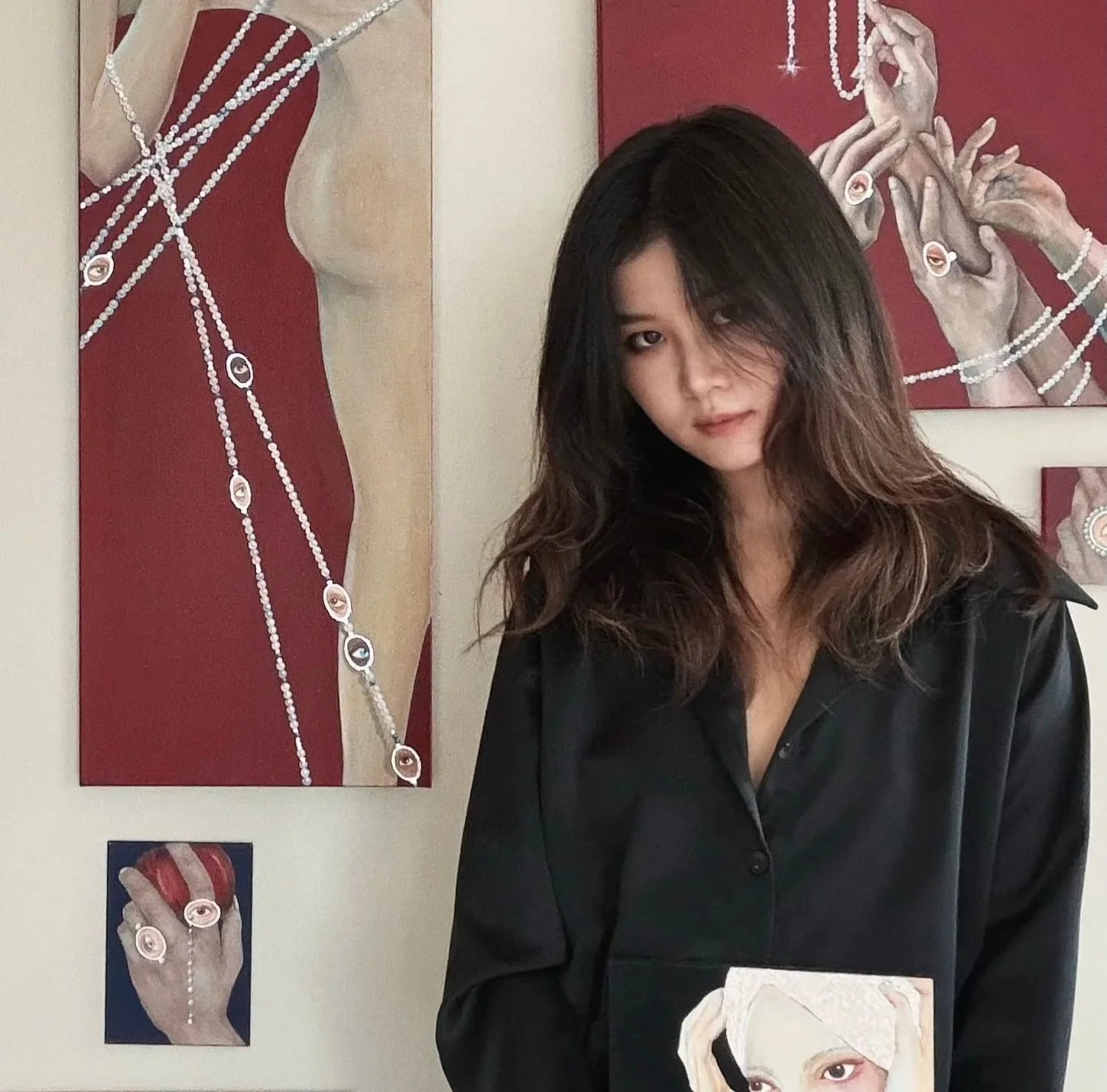 A woman with long dark wavy hair wearing a black shirt, standing in front of abstract art featuring hands and earrings made of beads and eye motifs, with smaller similar art pieces on the wall behind her.