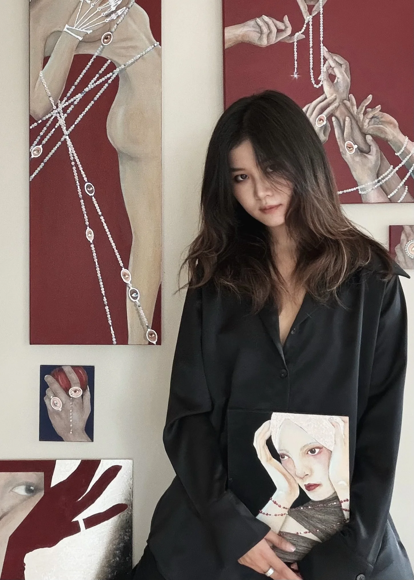 A woman with shoulder-length wavy hair, wearing a black button-up shirt, standing in front of colorful portraits of women, some with jewelry, on a gallery wall.