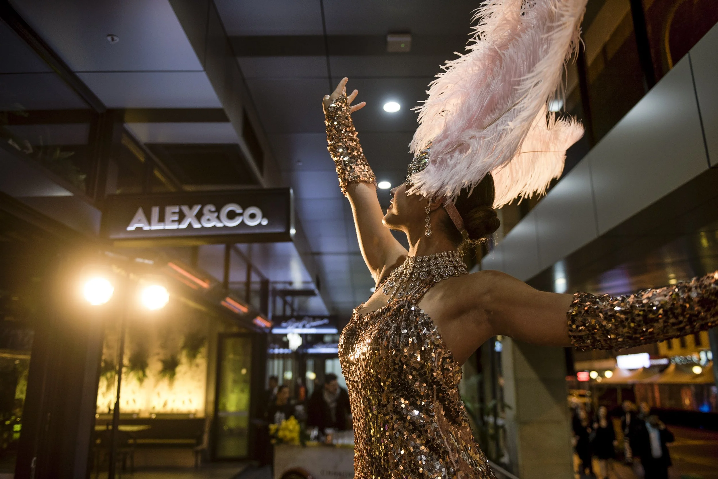 A woman dressed in a glamorous, sequined outfit, wearing large earrings, a statement necklace, and a feathered hat, performing or dancing with her arm extended in an indoor setting with restaurant signs and warm lighting.