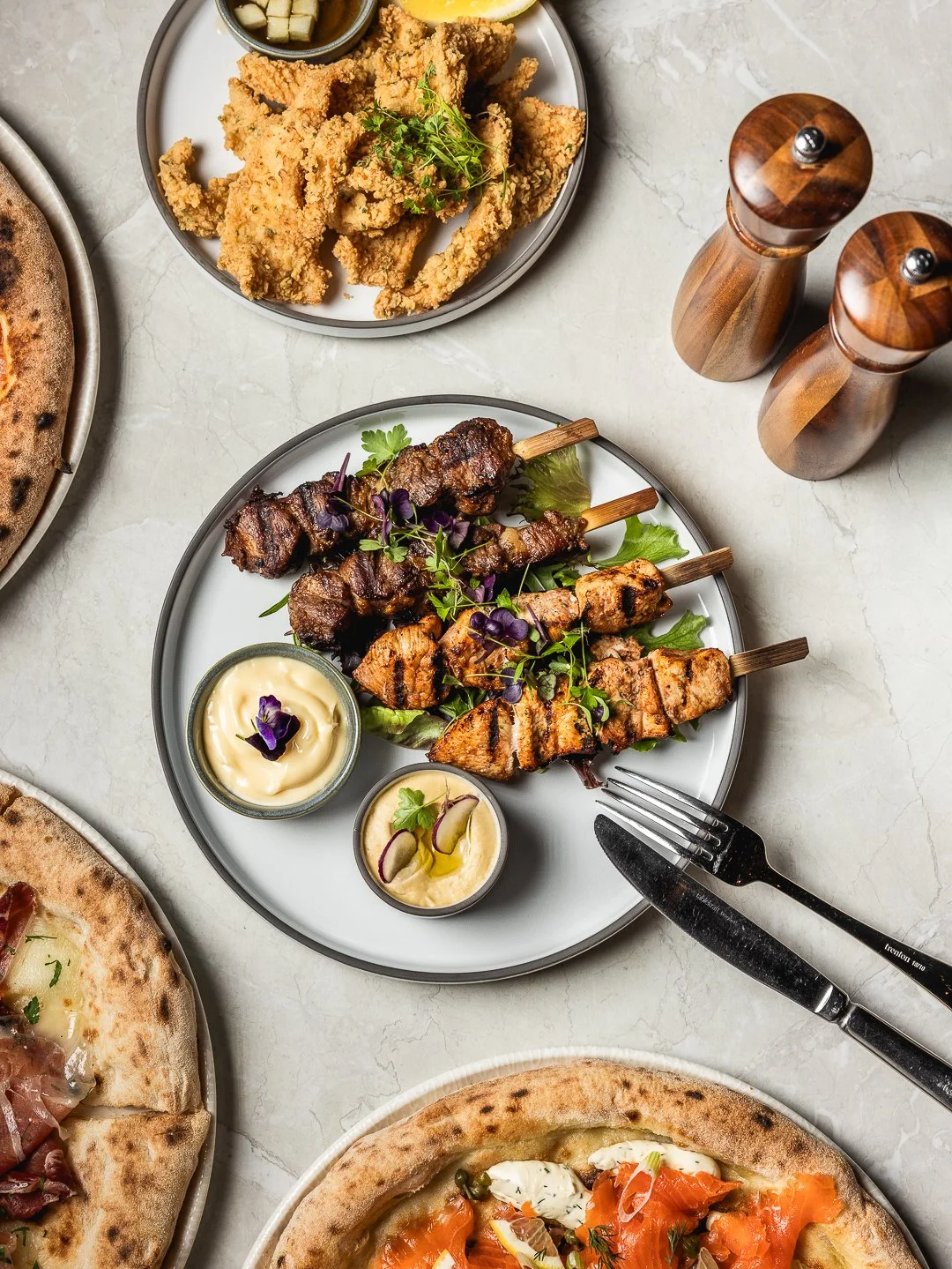 Plate of grilled meat skewers with green salad, accompanied by two types of sauce garnished with herbs. In the top section, a plate of fried chicken tenders with a dipping sauce and lemon wedge. Part of two pizzas, one with prosciutto and arugula, and the other with smoked salmon and herbs, are visible.