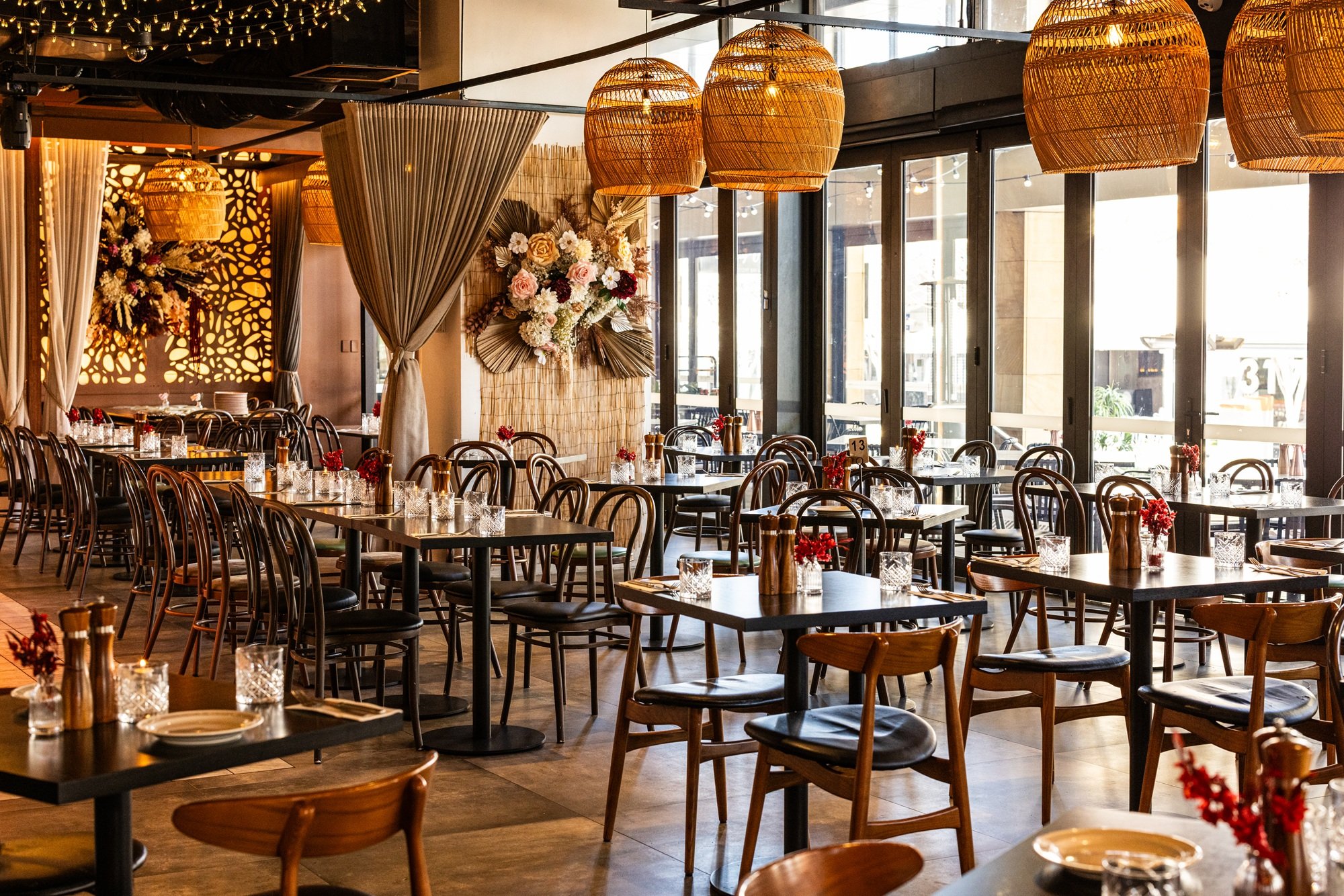 The restaurant has wooden chairs and tables with red flower centerpieces and glasses, decorated with warm hanging wicker lights and floral arrangements on the walls near large glass windows opening to the outside.