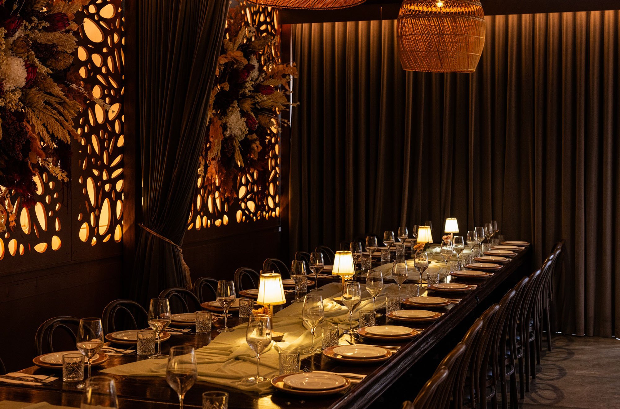 A long dining table set for a formal dinner in a dimly lit restaurant or event space, with wine glasses, plates, and small table lamps, illuminated by warm lighting and decorated with floral arrangements and a textured wall featuring illuminated cut-