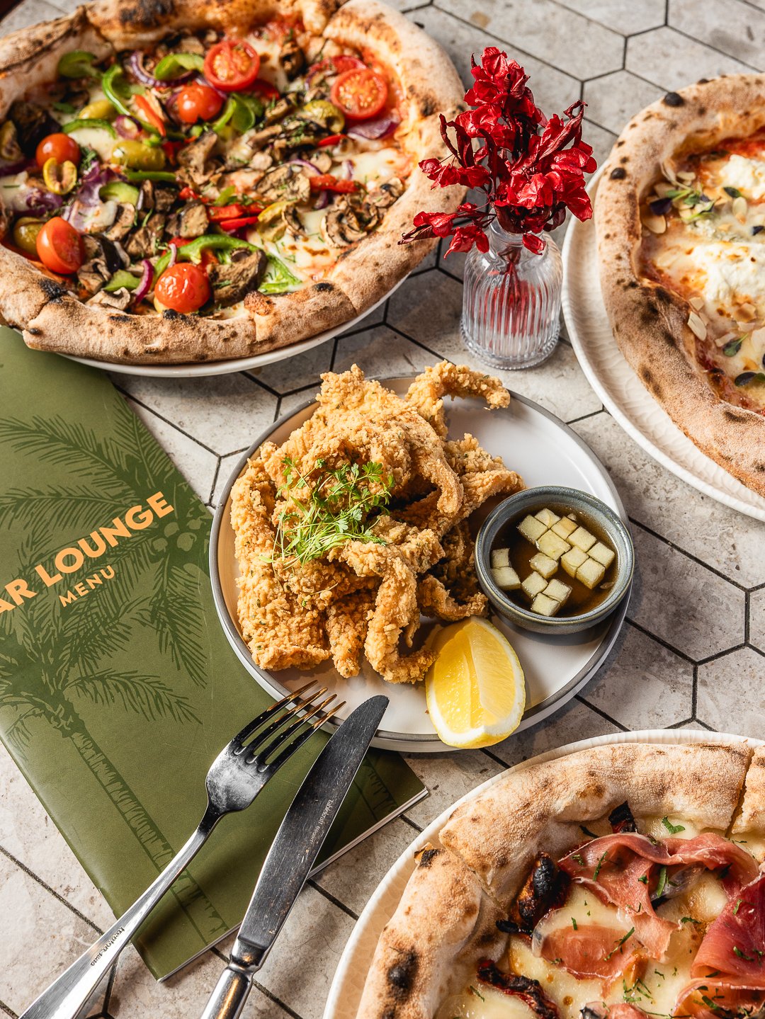 Pizza with cherry tomatoes, mushrooms, bell peppers, and onions on a white plate; fried chicken with lemon, dipping sauce, and garnish on a white plate; small vase with red leaves; green menu book titled 'Bar Lounge Menu' with a fork and knife on a t
