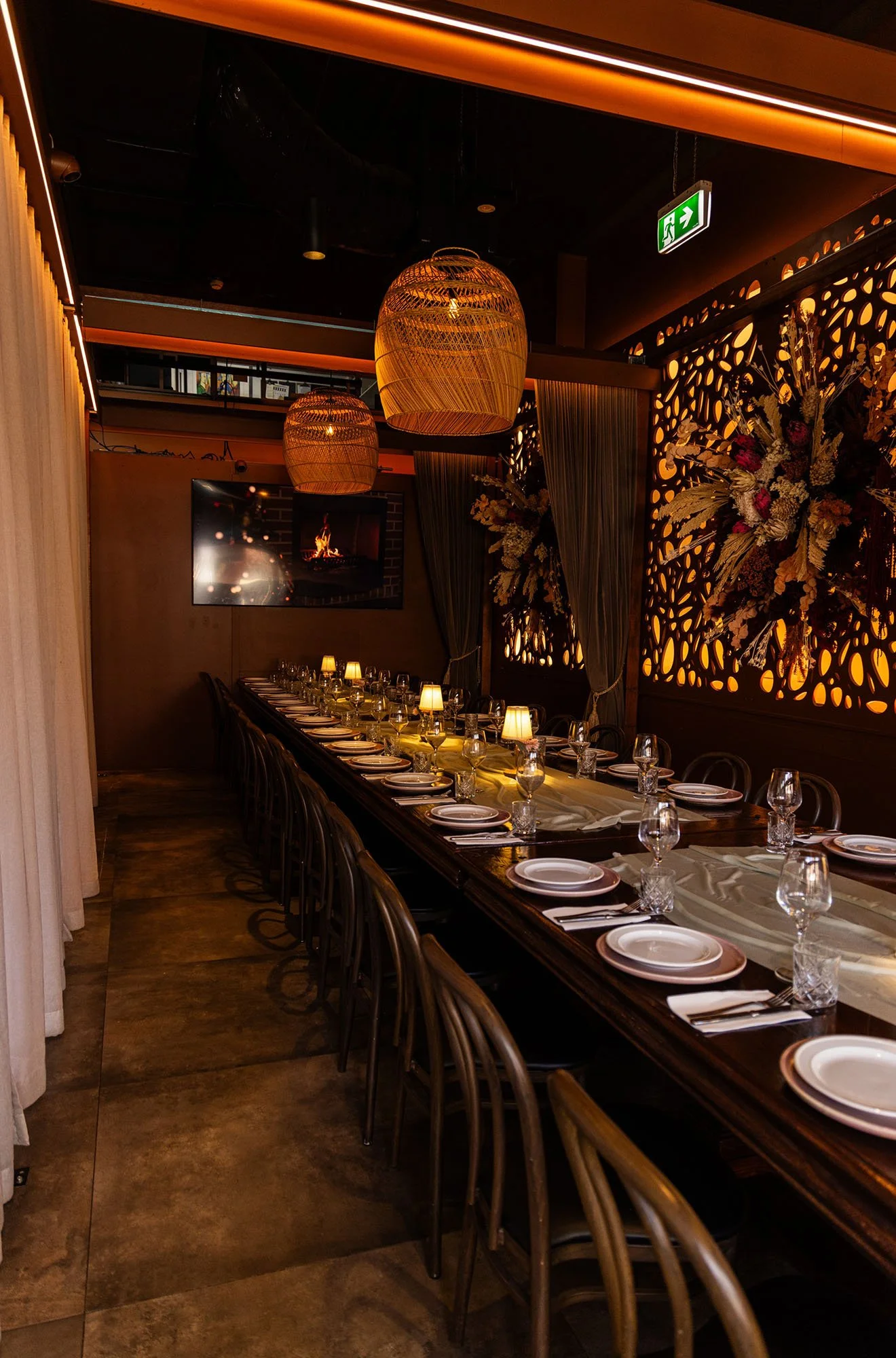 A long dining table set with plates, glasses, and napkins in a dimly lit restaurant with hanging woven pendant lights, floral wall decorations, and a television showing a fire, with curtains and an emergency exit sign.