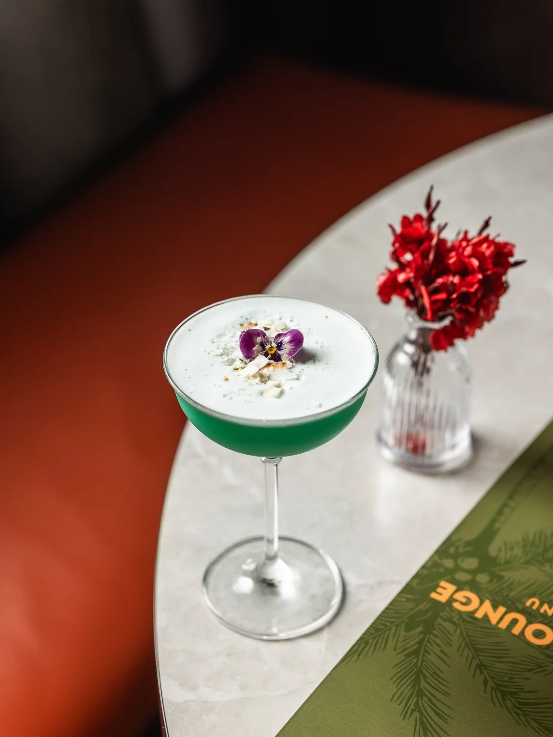 A green cocktail with white foam topped with an edible flower and nuts on a clear stemmed glass, placed on a white marble table next to a red flower in a clear vase.