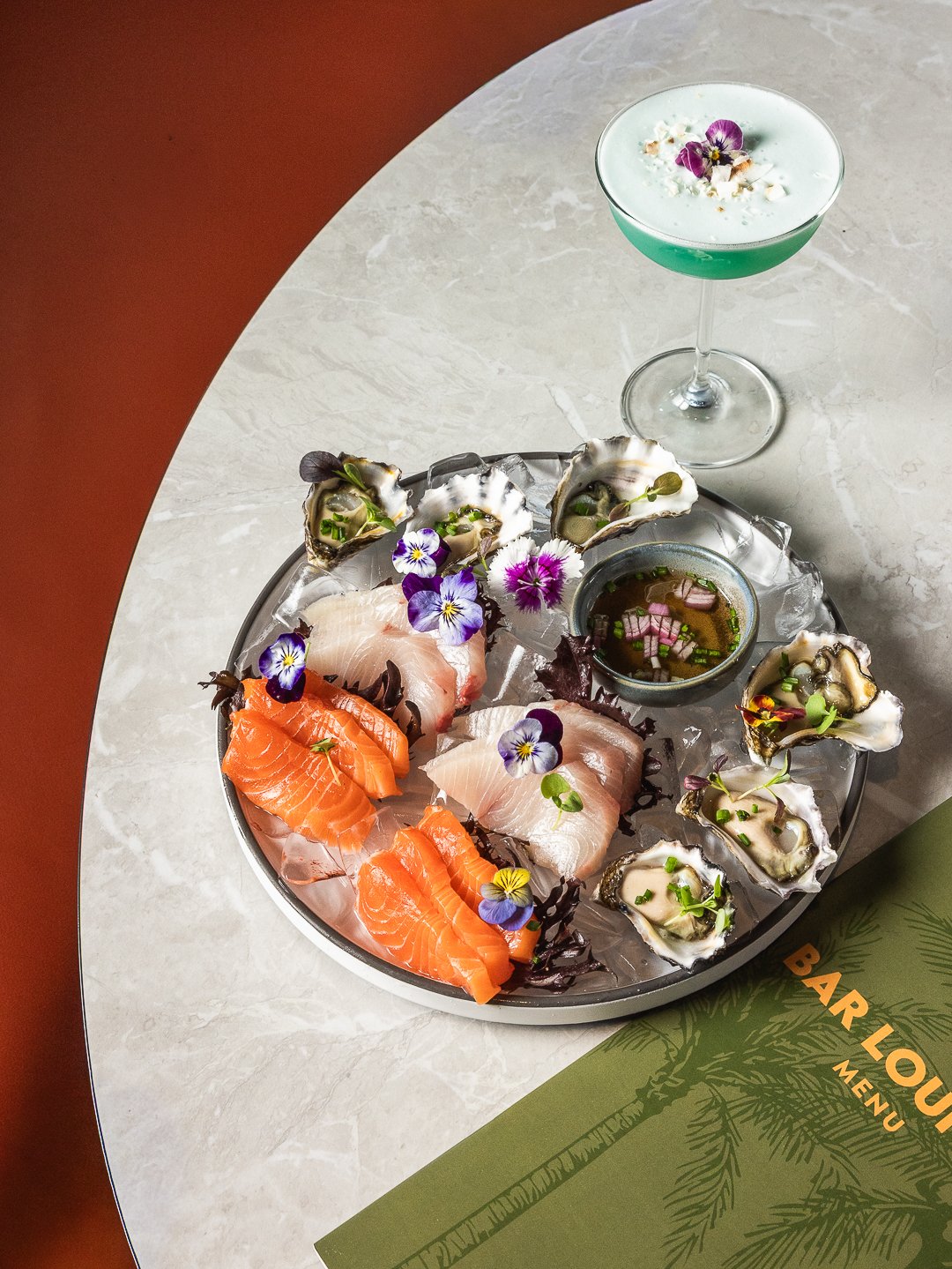 A round platter of sashimi including salmon, white fish, and oysters garnished with edible flowers on ice. A small bowl of soy sauce with chopped green onions and pink onions is in the center. An aqua-colored cocktail garnished with edible flowers is