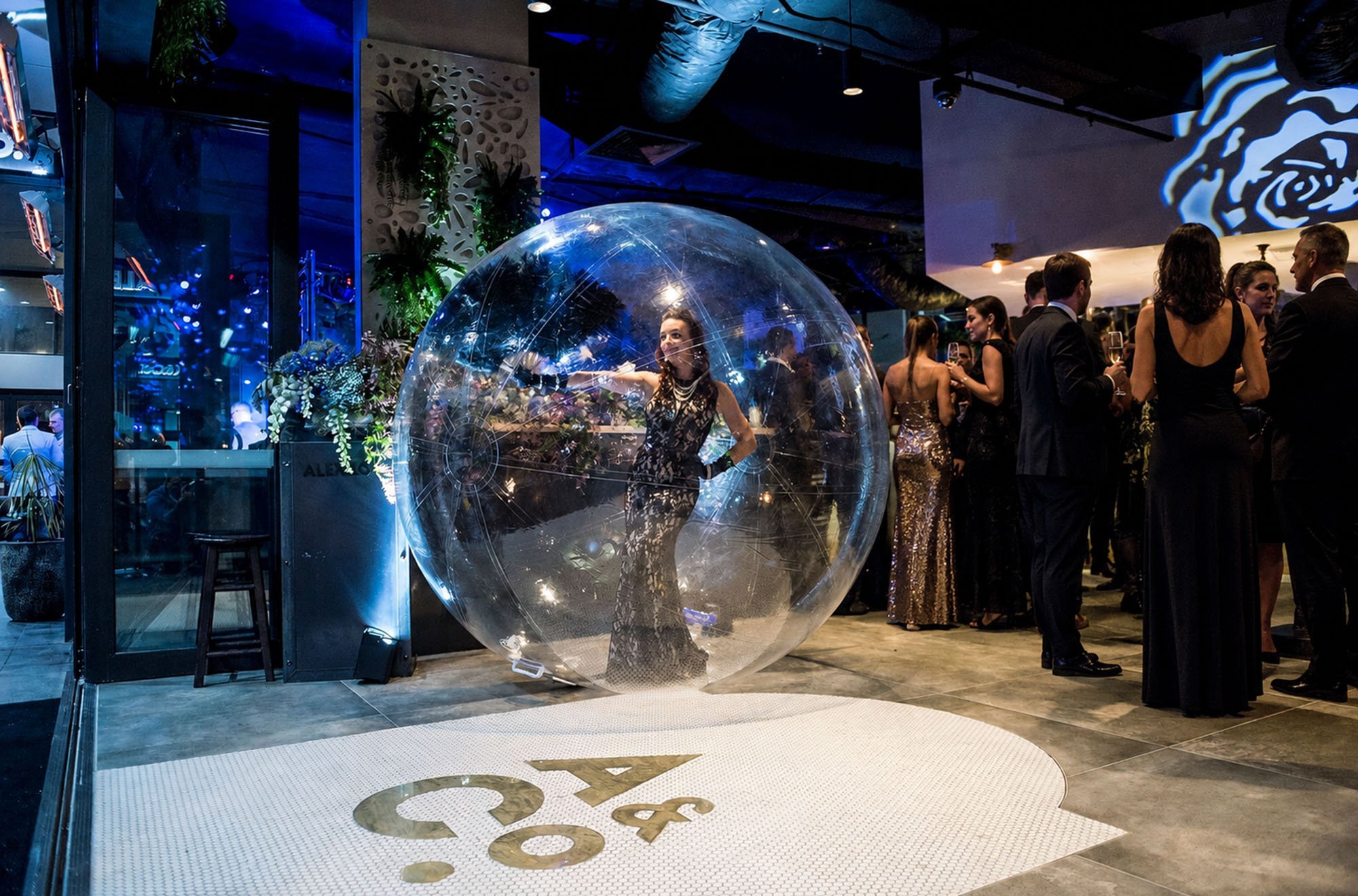People dressed in formal attire socializing at a nightclub or event with a modern, dark interior. A woman in a black dress stands inside a large, illuminated transparent sphere, while others mingle nearby.