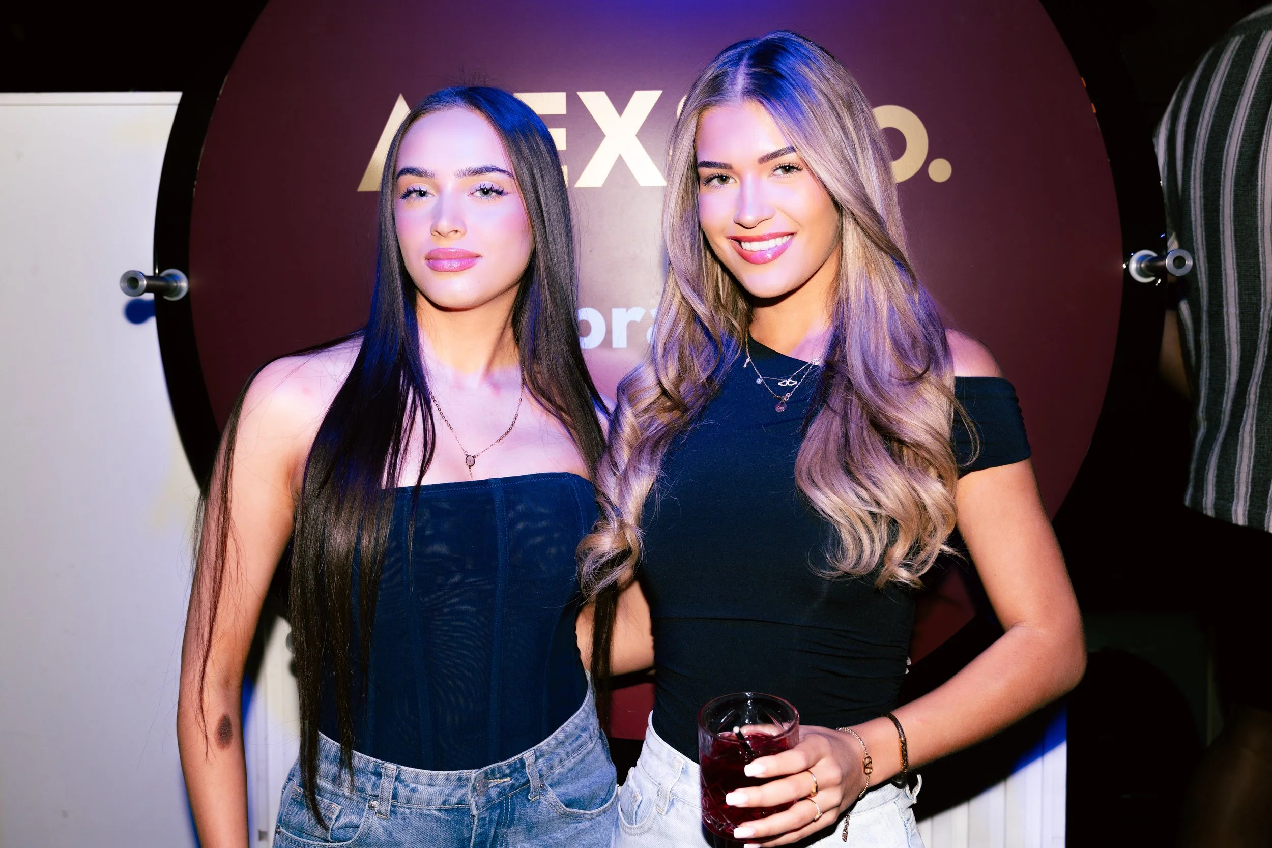 Two young women standing close together at a social event, both smiling and holding drinks. One woman has long dark hair and the other has long blond hair. There is a dark background with the partial text 'ALEX' visible behind them.