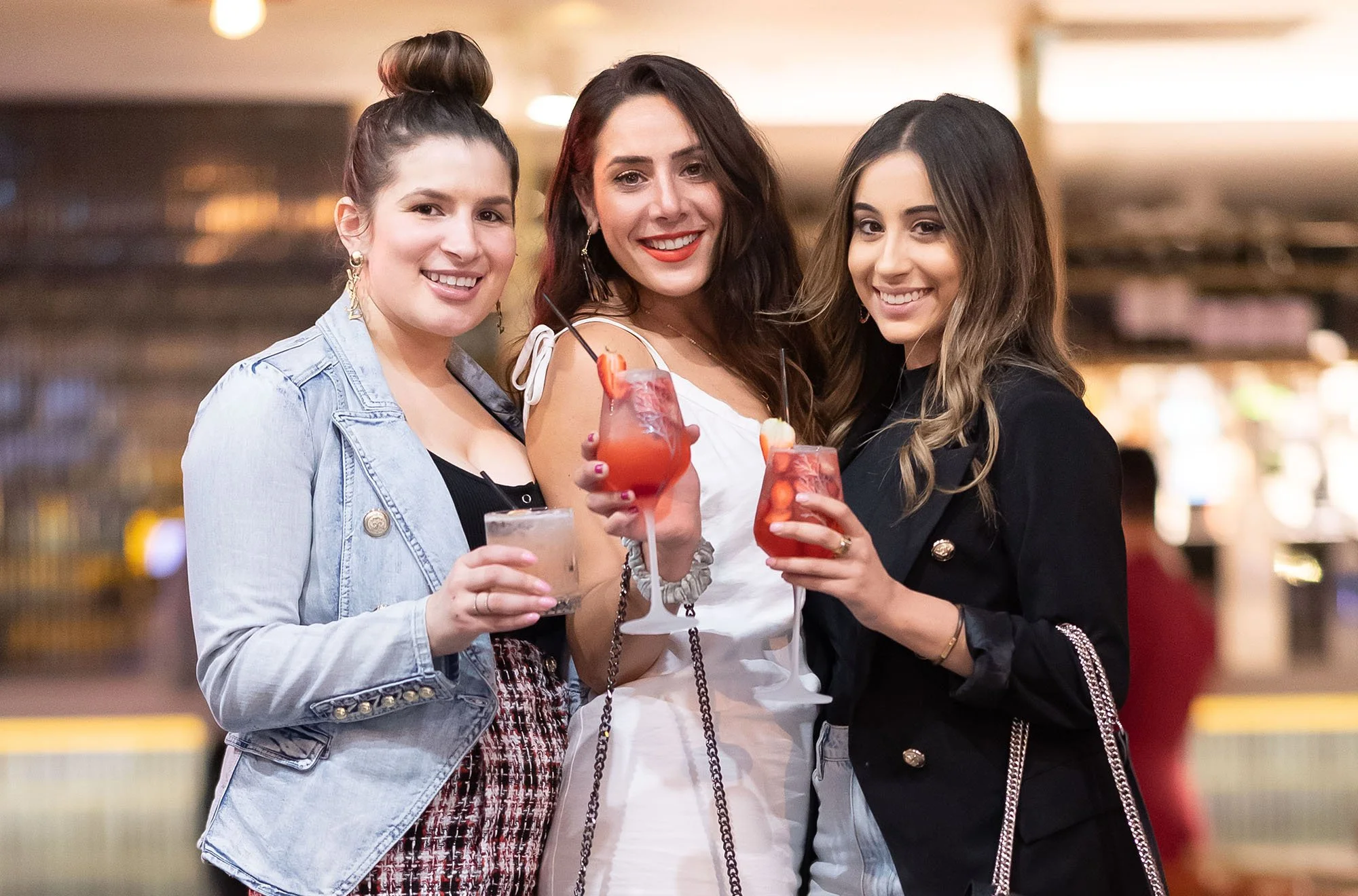 Three women smiling and holding colorful cocktails in a lively indoor setting.