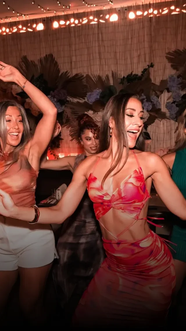 Group of women dancing and having fun at a party or celebration indoors with string lights and decorative foliage on the wall behind them.