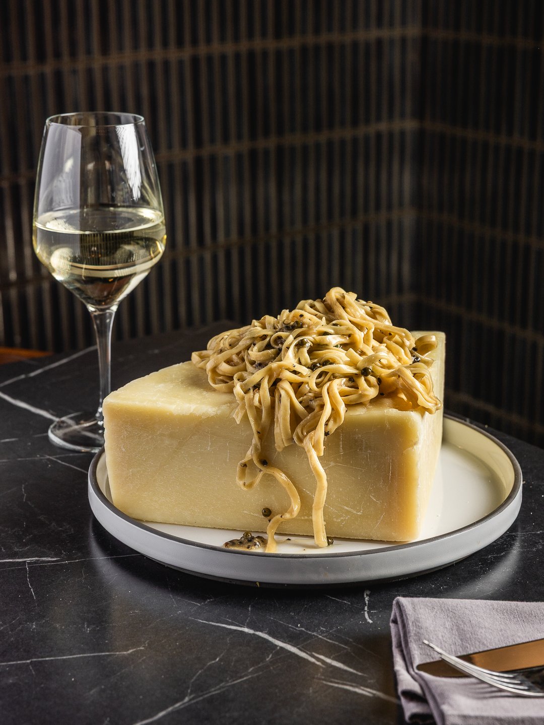 A block of cheese topped with cooked noodles and black peppercorns, accompanied by a glass of white wine, on a black marble table with a gray napkin and metal fork.