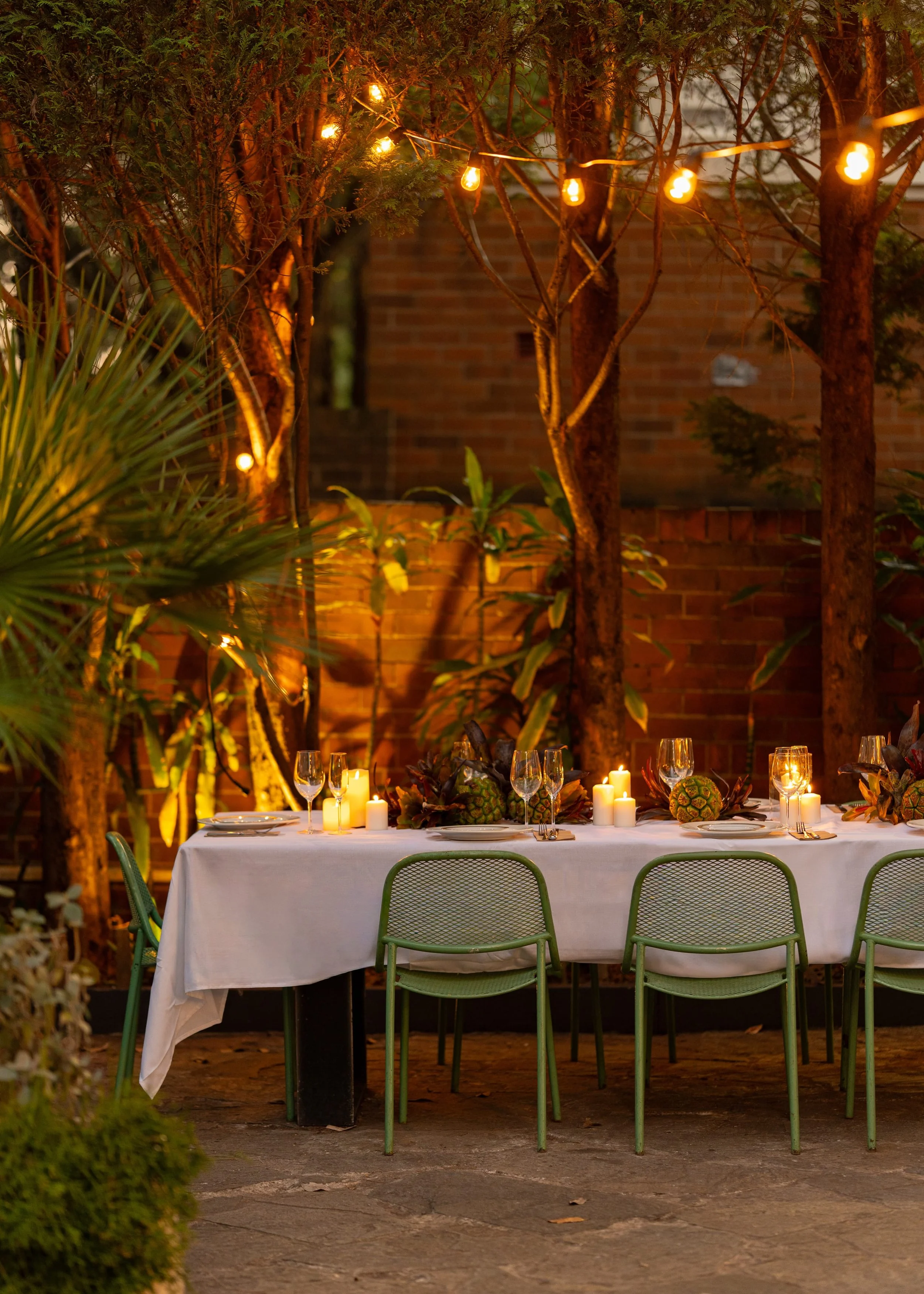 An outdoor dining table decorated with candles and greenery, set for a meal, under string lights, with trees and a brick wall in the background during evening.