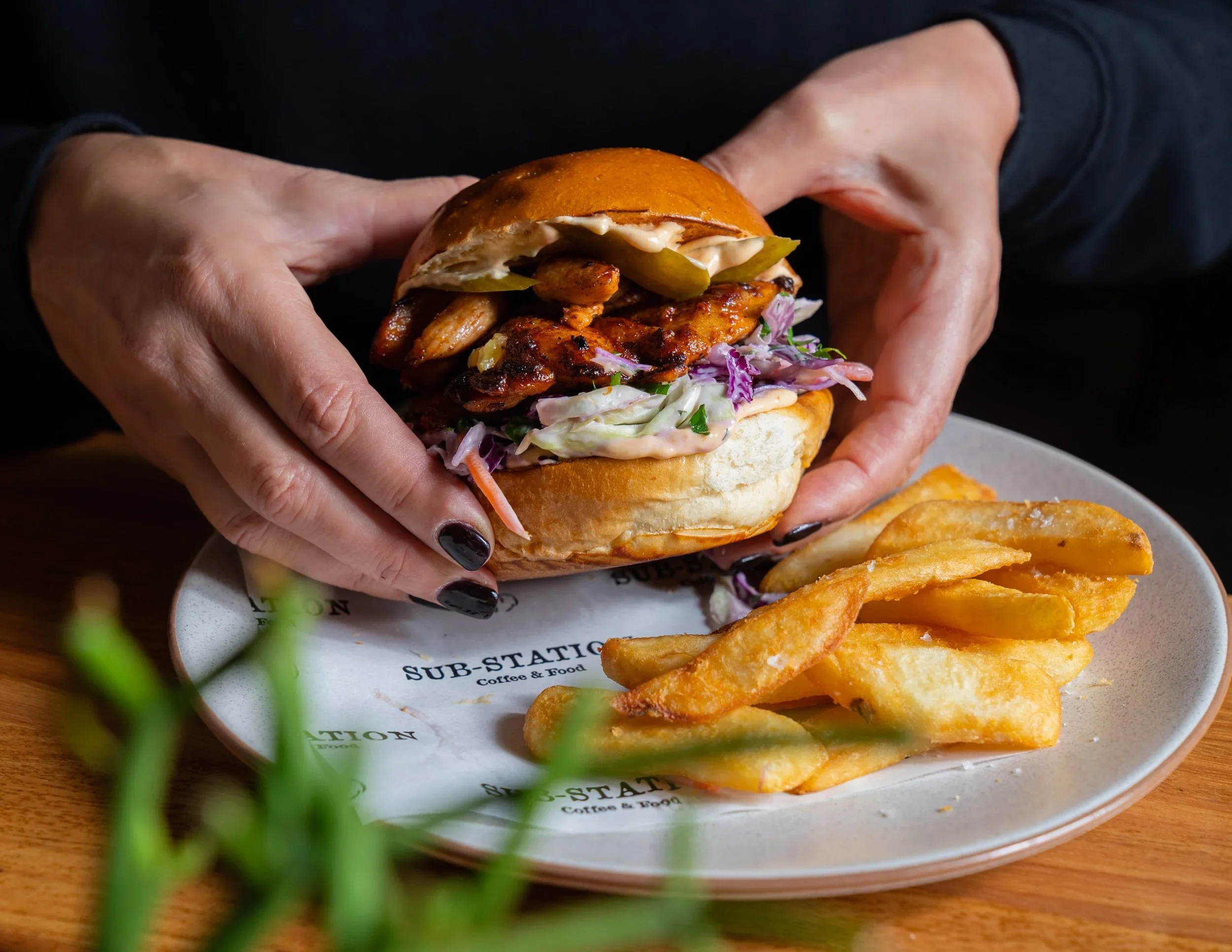 A person holding a toasted sandwich with grilled chicken, lettuce, pickles, coleslaw, and mayonnaise inside a bun. A plate with fried potato wedges is in the foreground, on a wooden table, with a blurred green plant partially visible.