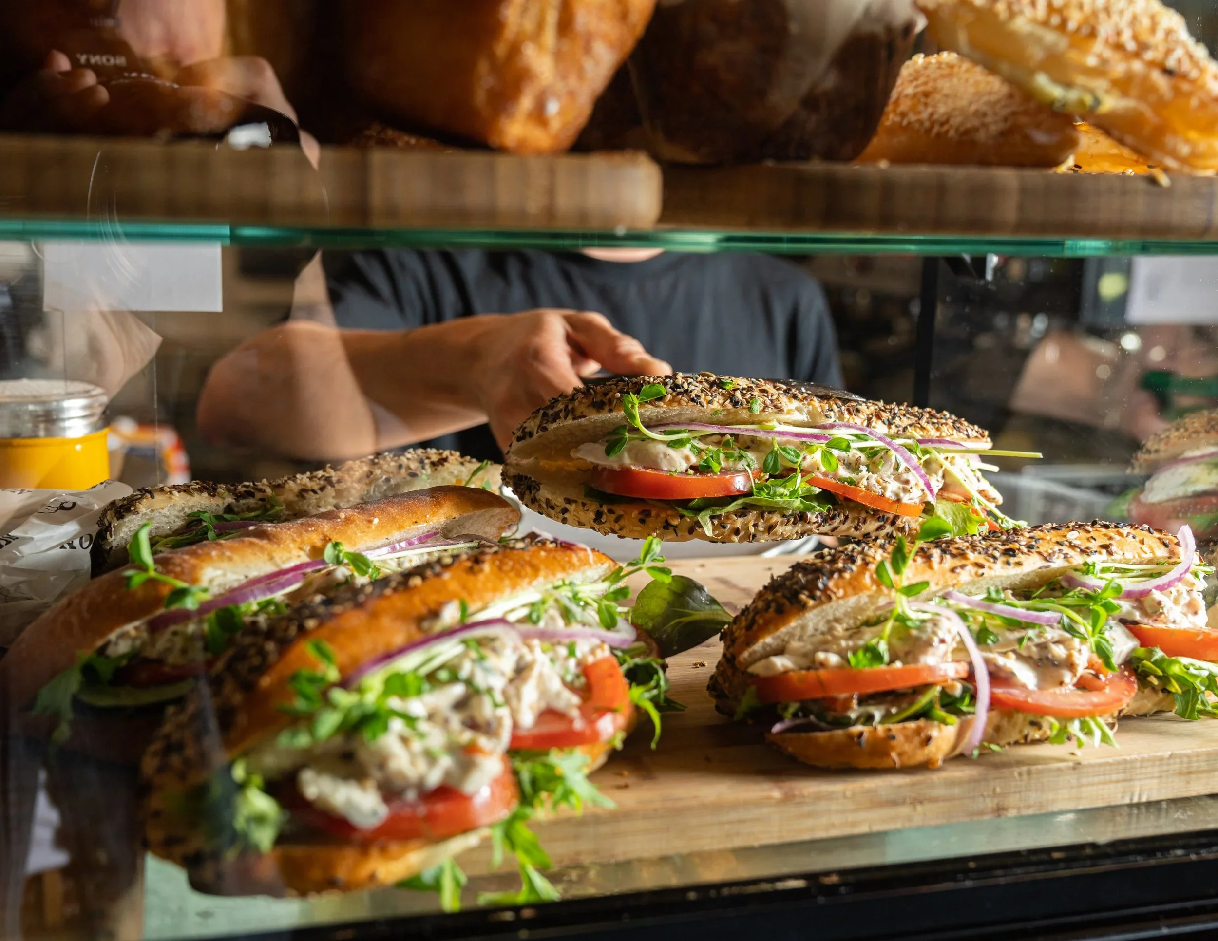 A bakery display case filled with sandwiches topped with sesame seeds, fresh vegetables, and herbs, with a person behind the case preparing more sandwiches.