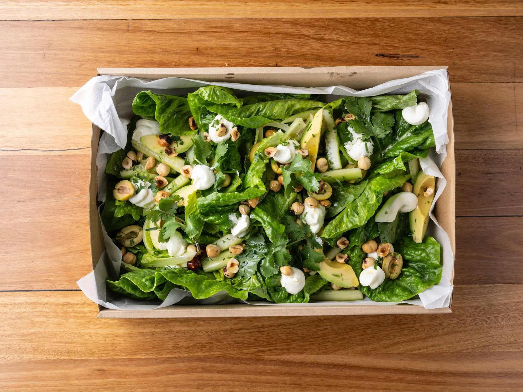 A rectangular cardboard box lined with parchment paper, filled with a fresh green salad including lettuce, avocado slices, mozzarella balls, hazelnuts, and chopped herbs, on a wooden surface.