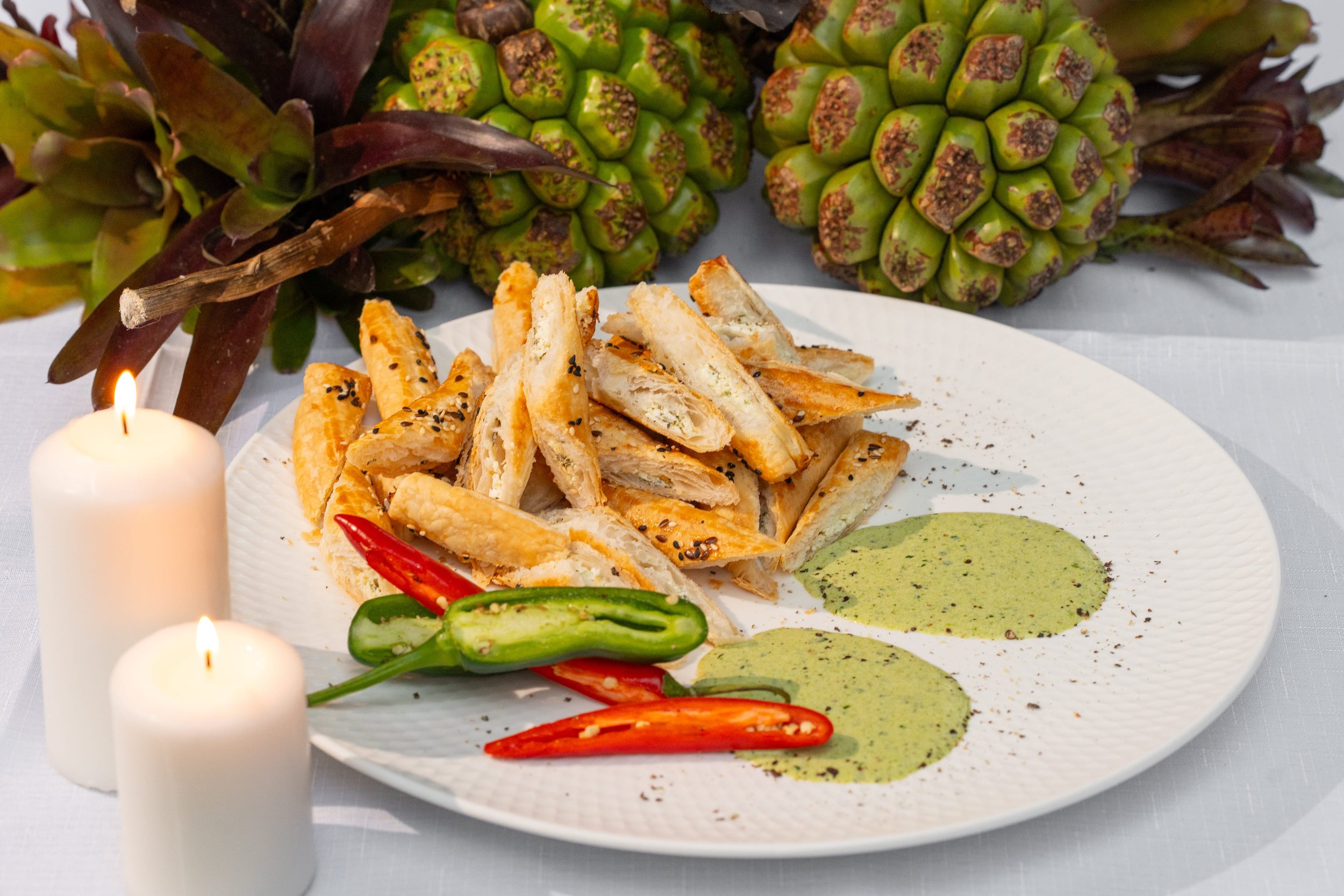 Plate of sliced chicken with spices, colorful peppers, green dipping sauce, and fresh pineapples in the background, with candles around.