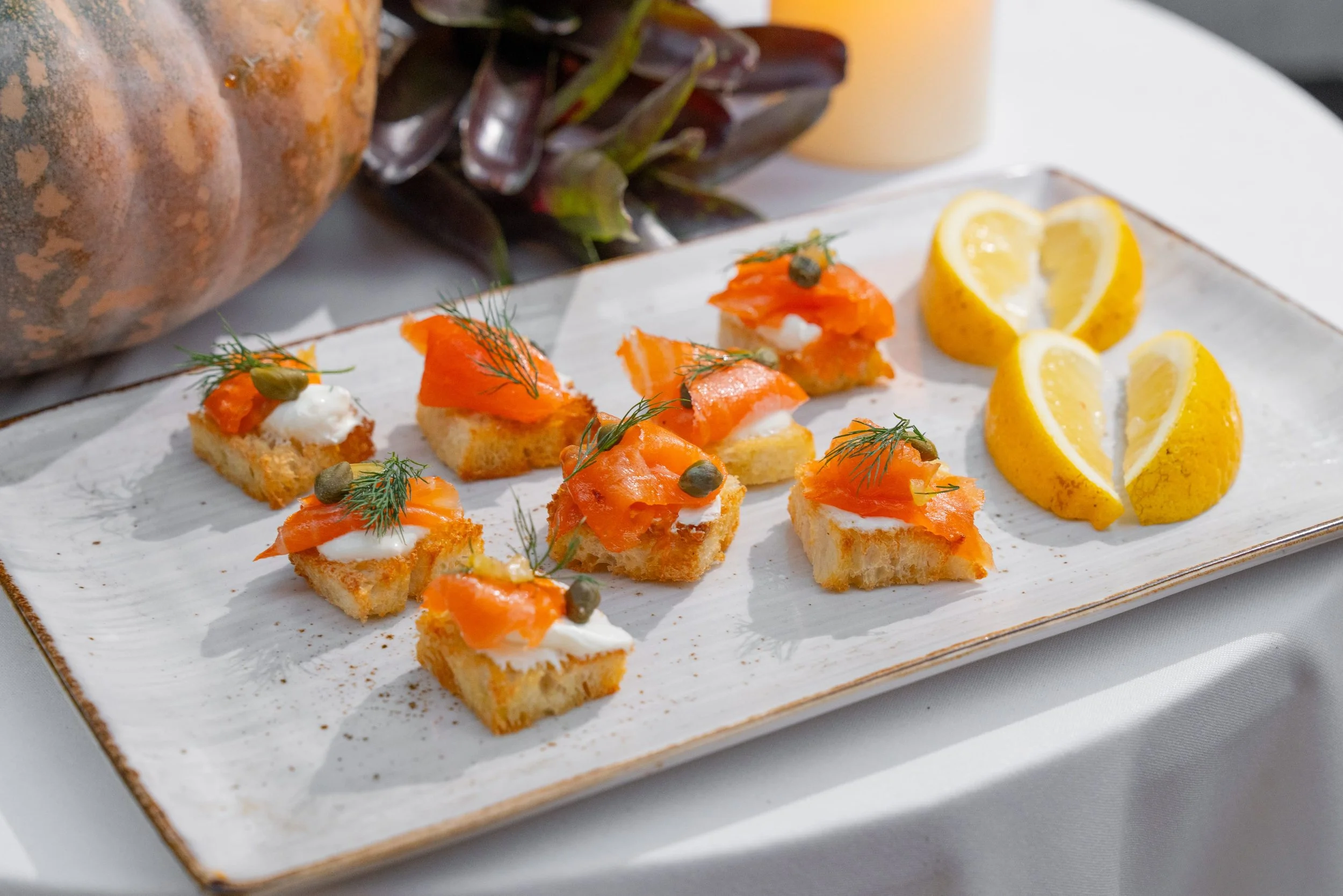 Canapés with smoked salmon, dill, capers, and cream cheese on slices of baguette. Lemon wedges on the side, on a rectangular white ceramic platter.