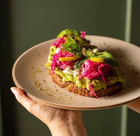 Open-faced avocado toast topped with pickled pink onions, herbs, and sliced yellow peppers on a beige plate