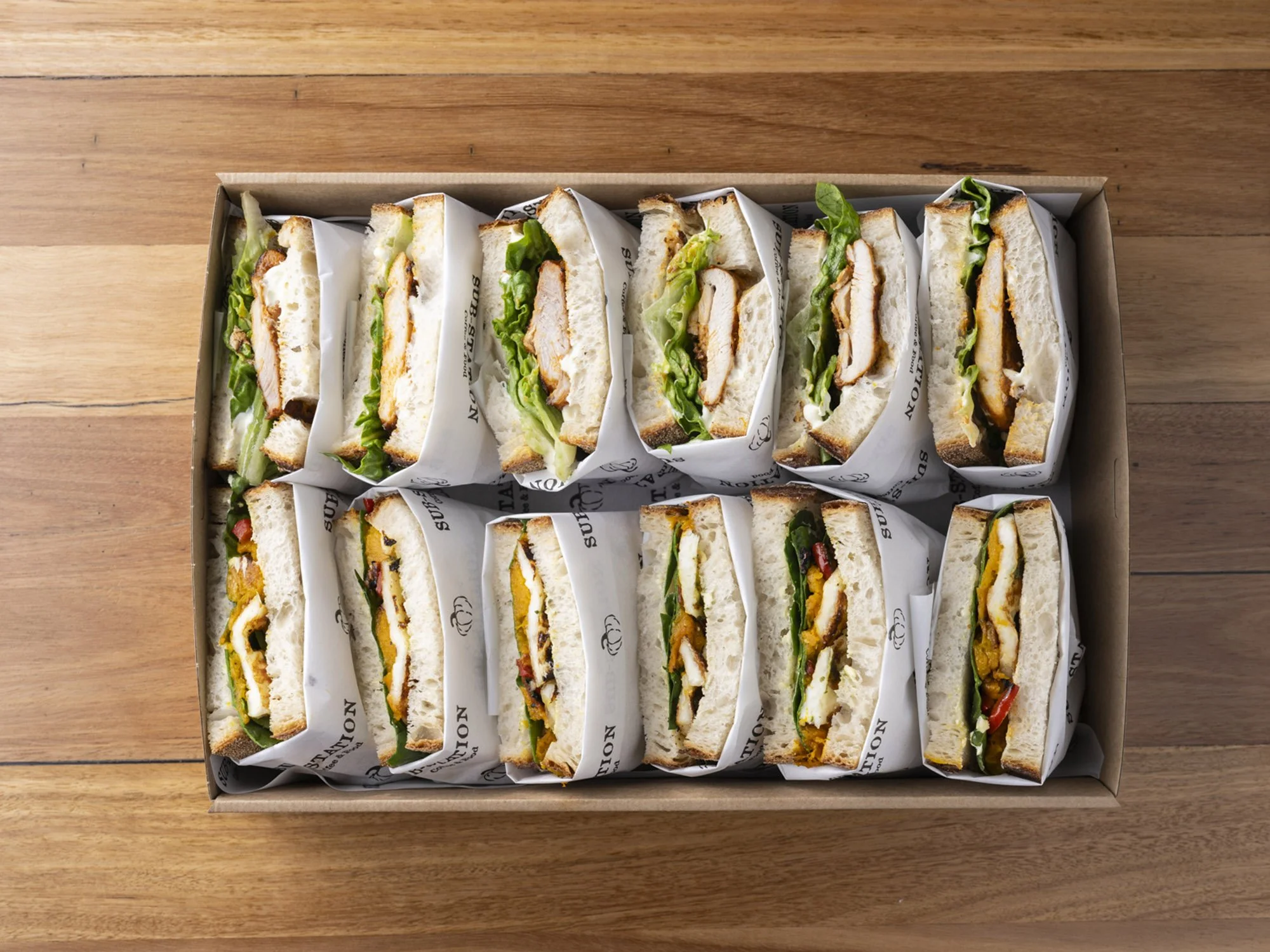 A box of assorted sandwiches with bread, lettuce, chicken, and other ingredients, wrapped in paper with 'SUBWAY' printed on it, placed on a wooden surface.
