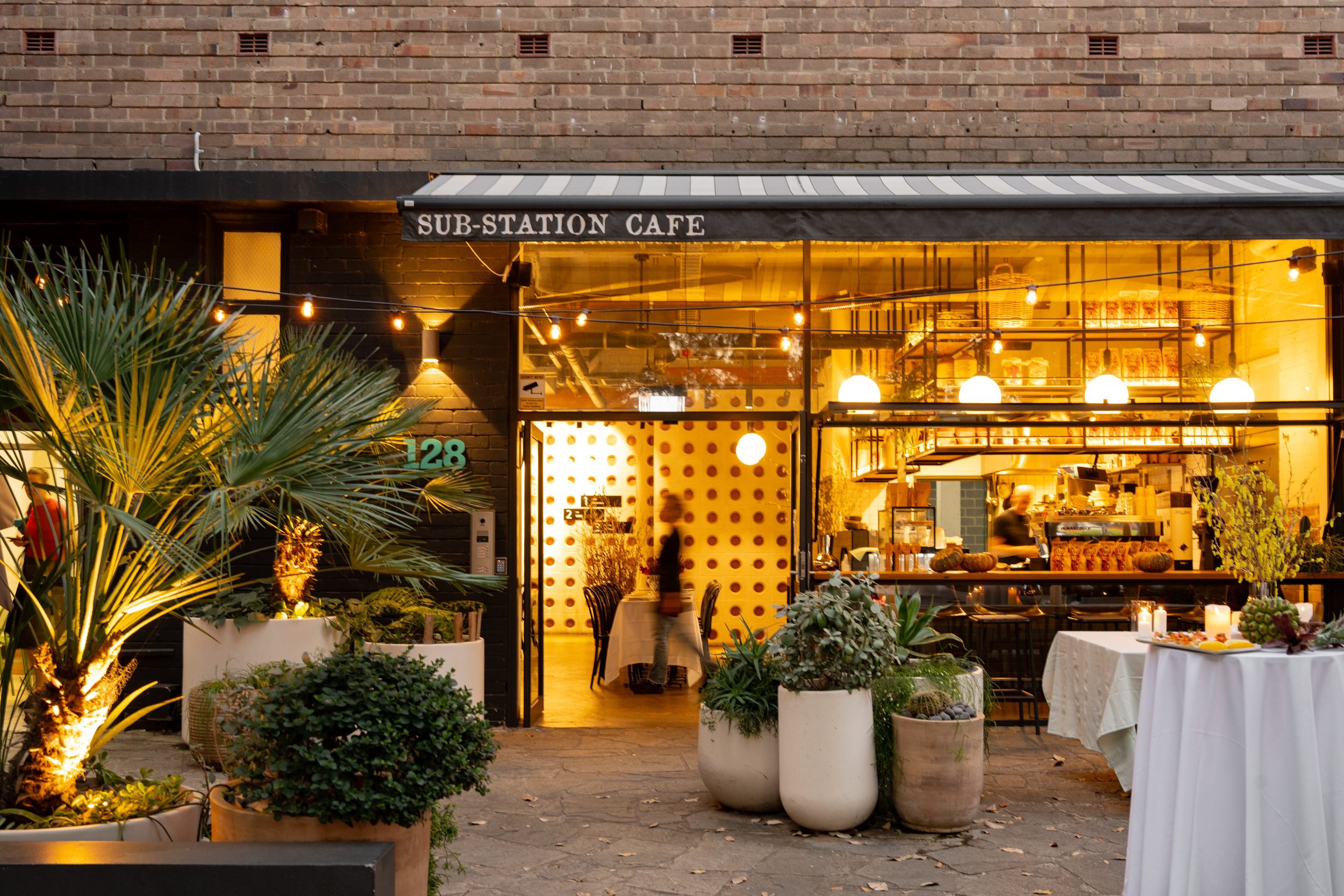 Night view of a cozy restaurant named SUB-STATION CAFE with warm yellow lighting. The storefront has large glass windows, outdoor seating with tables and chairs, potted plants, and decorative string lights.