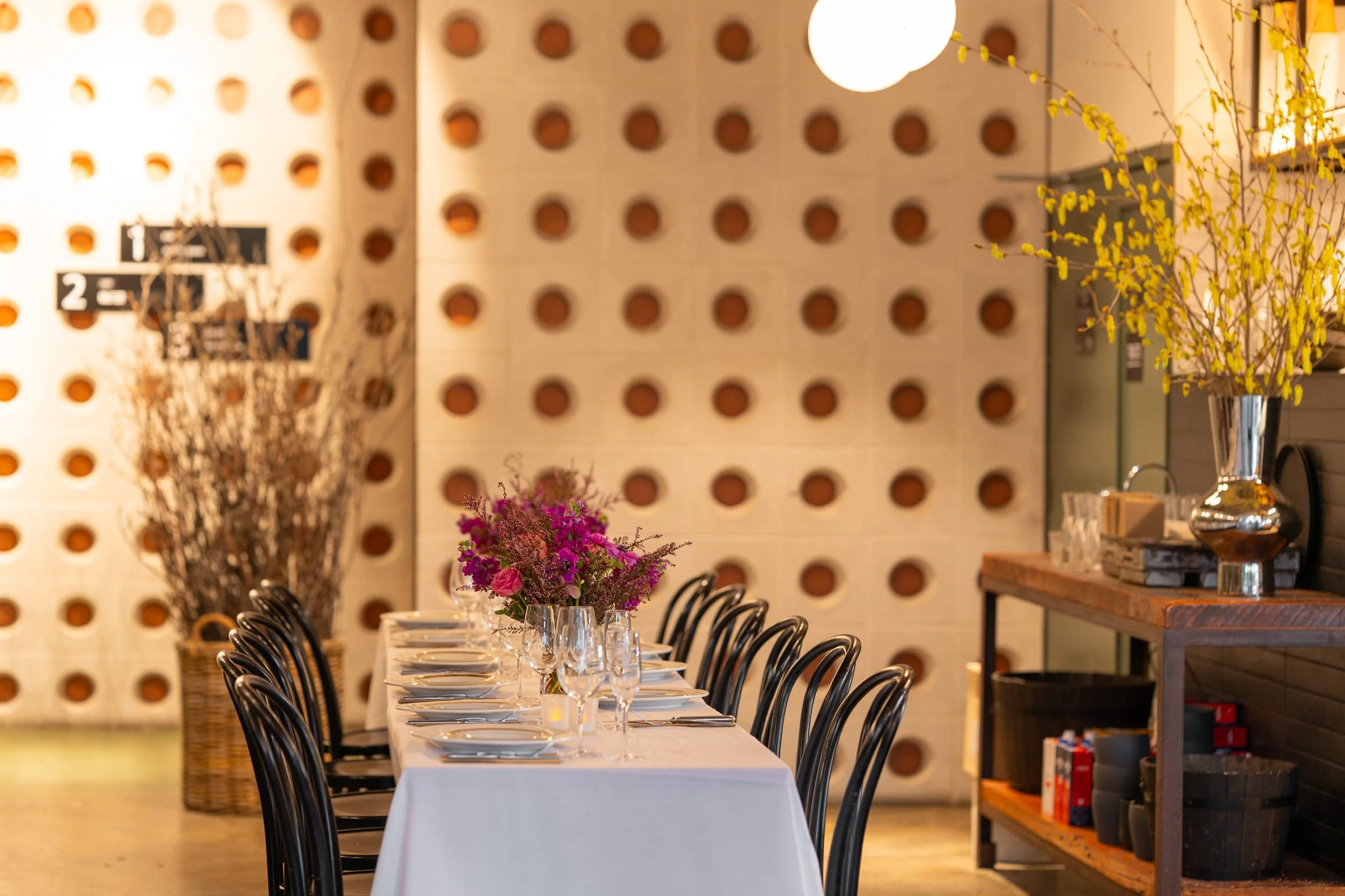 A long dining table set with white tablecloths, glassware, and floral centerpieces, positioned in a modern interior with a wall featuring large round holes and various decorative elements, including a tall vase with yellow flowers and a wooden shelf.