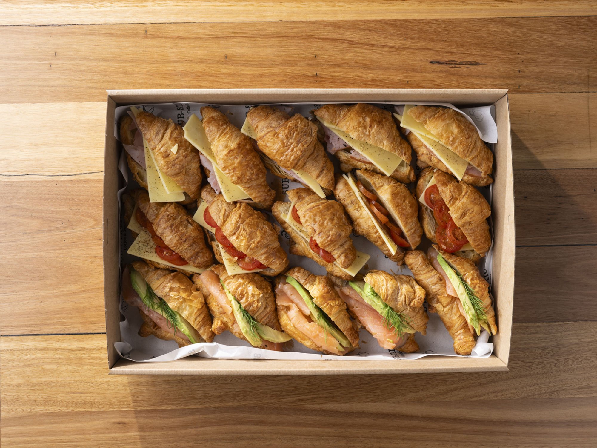 A box of assorted croissant sandwiches with cheese, tomato, ham, salmon, and vegetables on a wooden table.