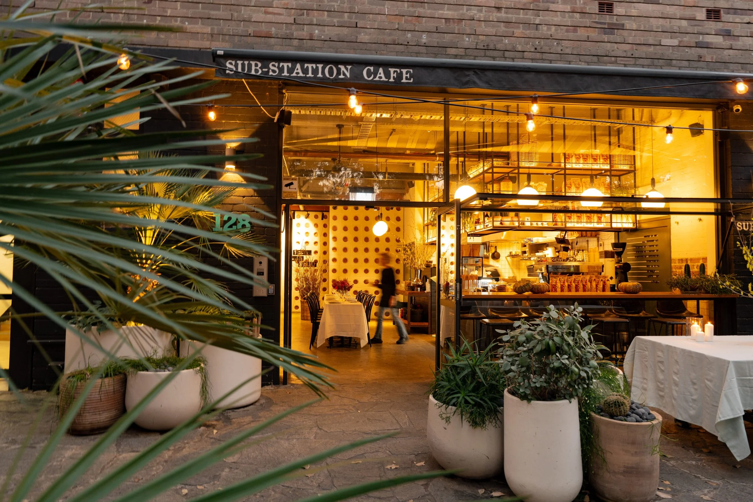 Exterior view of Sub-Station Cafe with large windows, warm yellow lighting, potted plants outside, and tables set for dining.