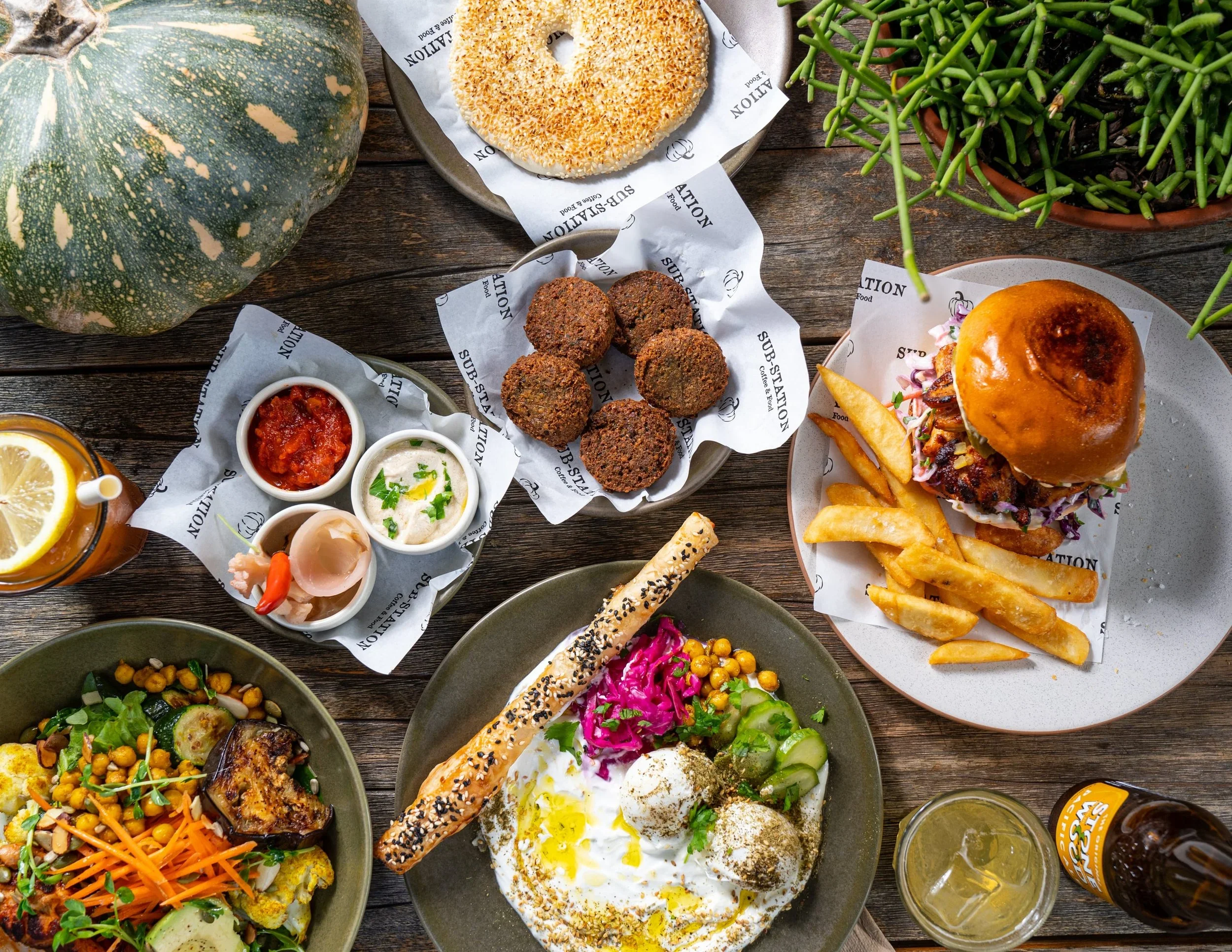 Assorted foods on a wooden table including a large green pumpkin, a sesame seed bagel, falafel balls, a burger with fries, and various salads and dips.