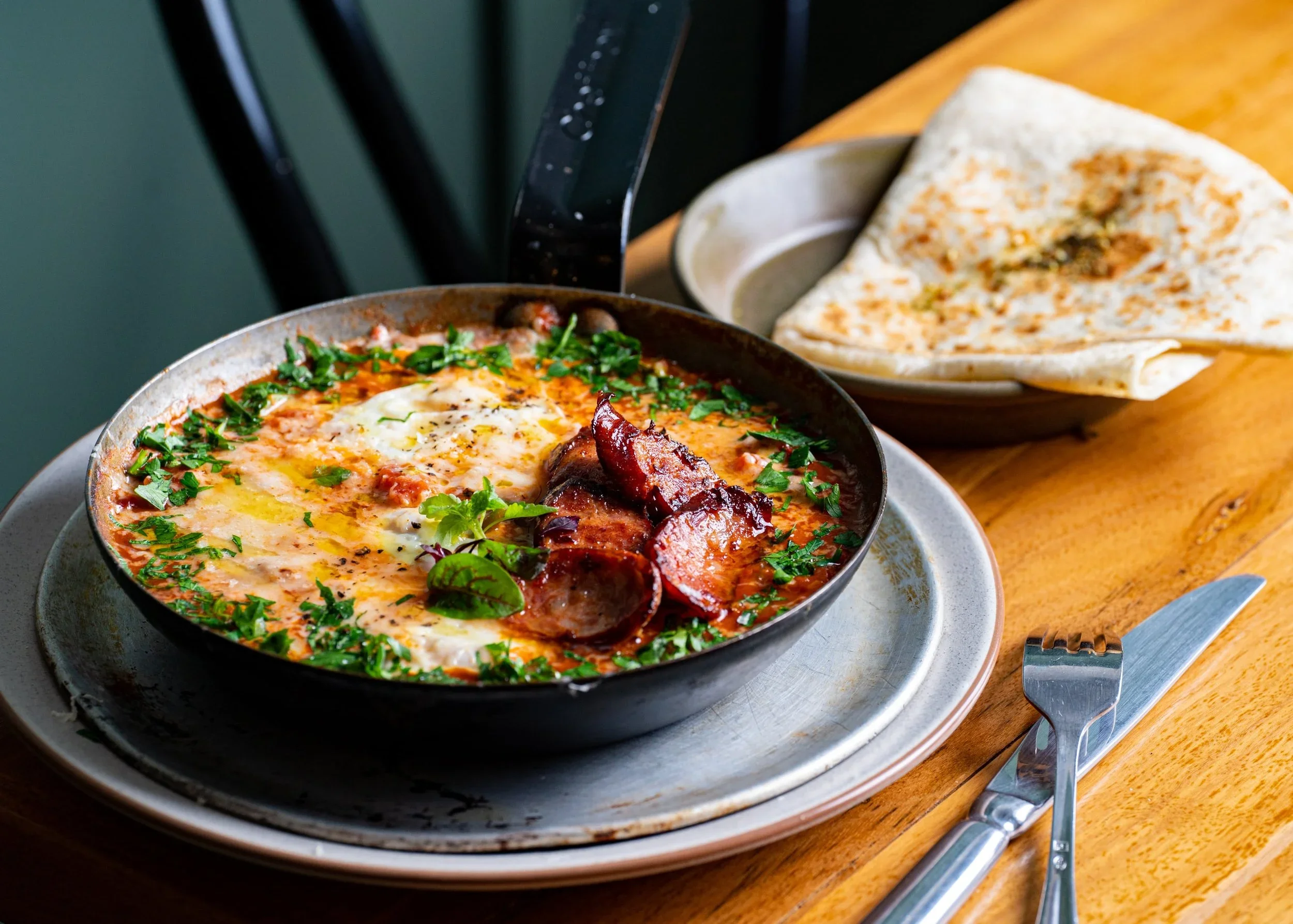 A skillet of baked eggs topped with fresh herbs and cooked tomatoes, with a plate of flatbread in the background on a wooden table.