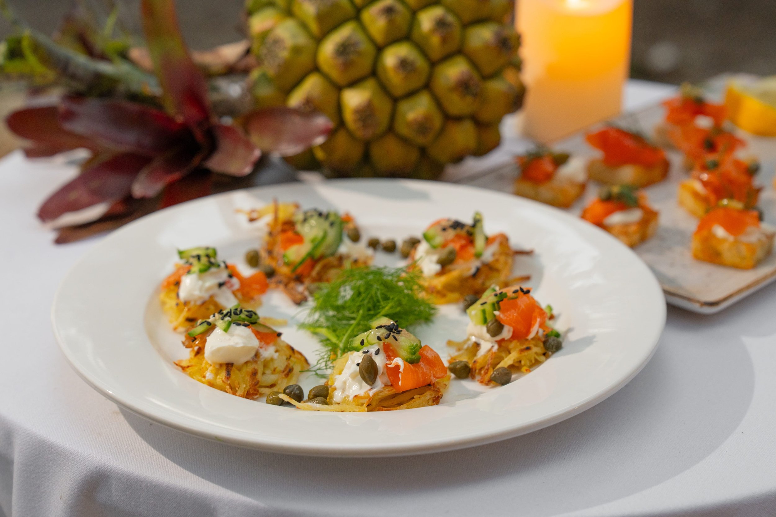A white plate with Asian-style rice topped with smoked salmon, cucumber, capers, and black sesame seeds, garnished with fresh dill. In the background, there is a whole pineapple, some orange-colored finger foods, and a glass of orange juice.