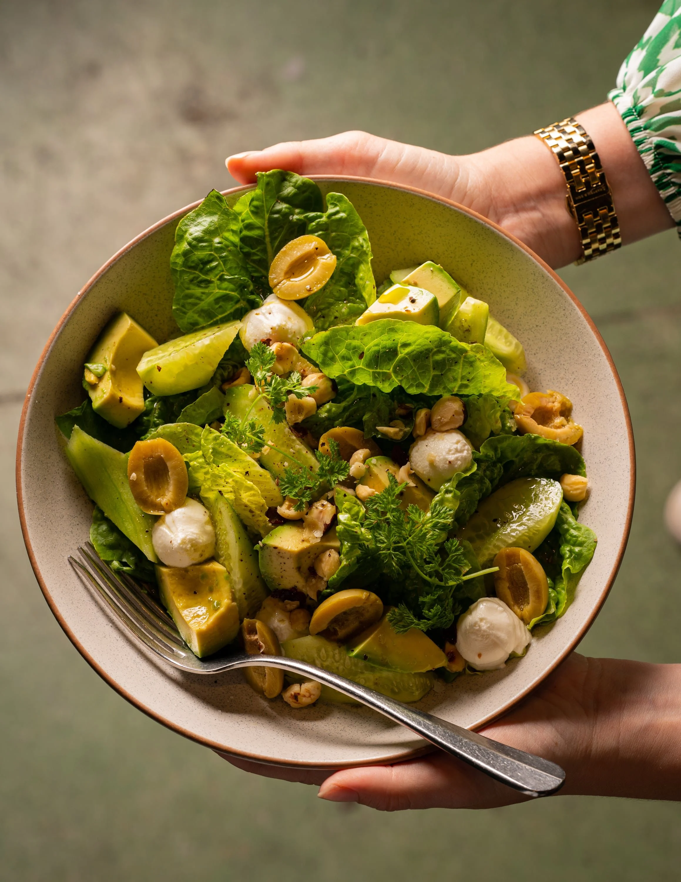 A hand holding a bowl of salad with lettuce, avocado, cheese, olives, and herbs, with a fork resting on the edge of the bowl.