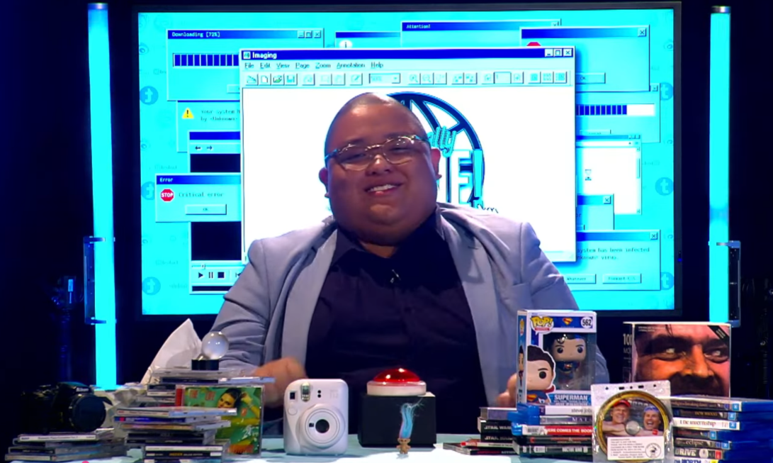 A man in glasses and a gray blazer smiling at a desk cluttered with books, DVDs, a camera, and Funko Pop figures, with a large computer screen displaying overlapping old Windows error and download windows in the background.