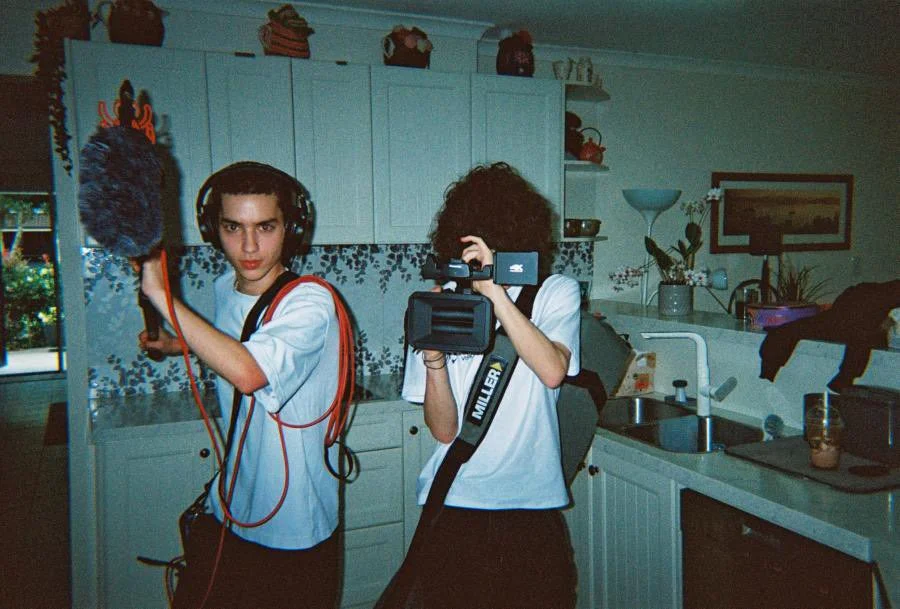 Two young men filming in a kitchen, one holding a camera and the other holding a microphone with a windscreen, wearing headphones and casual clothing.