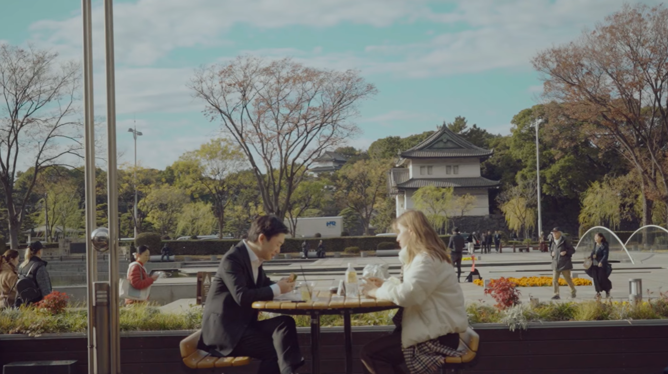 Two women sit across from each other at an outdoor table, using their phones, with a cityscape and autumn trees in the background.