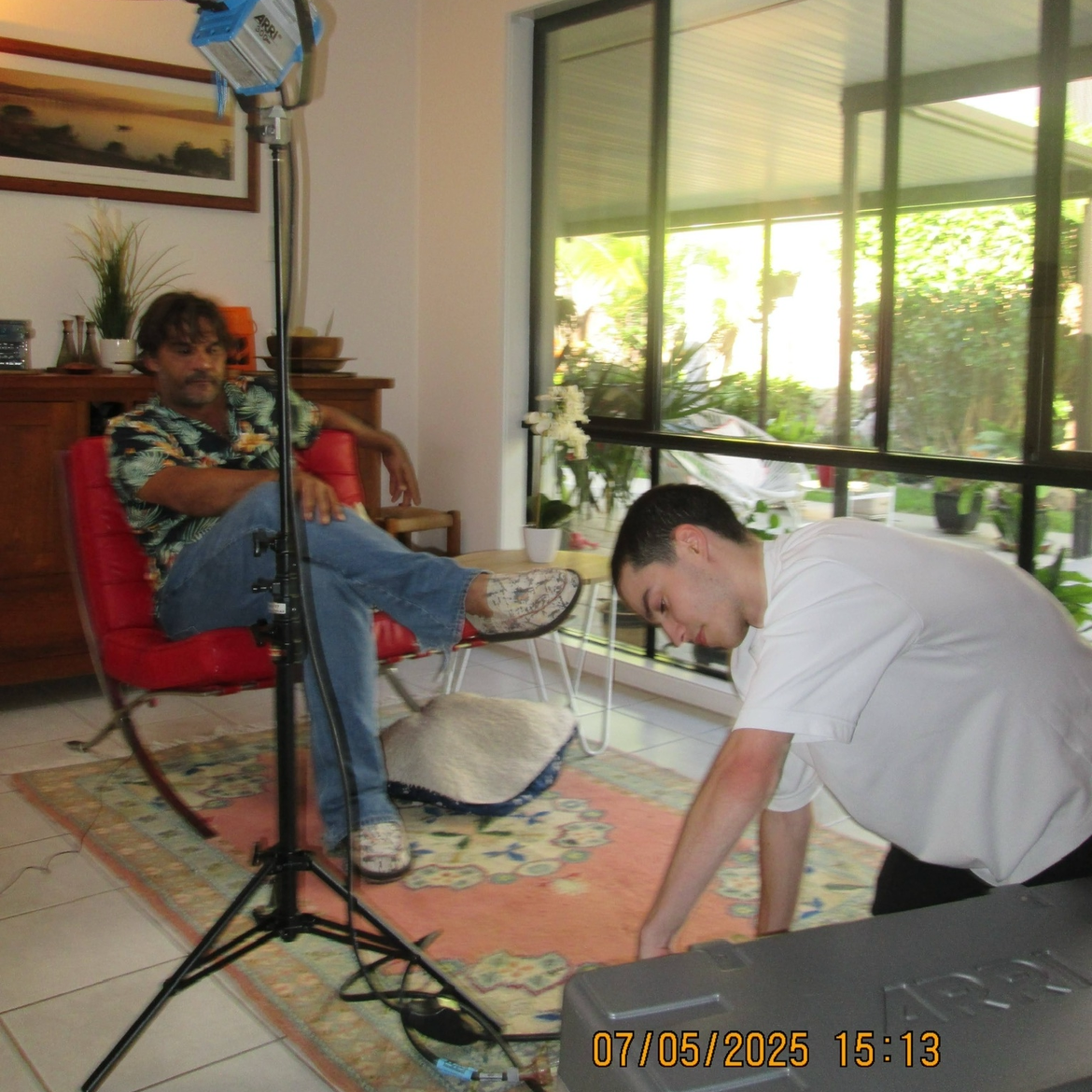 A man sitting in a red chair with one leg crossed over the other, wearing a patterned shirt, jeans, and sneakers. A younger man in a white shirt is leaning forward on the floor. The scene is set in a bright room with large windows, a wooden sideboard, potted plants, and a colorful rug.