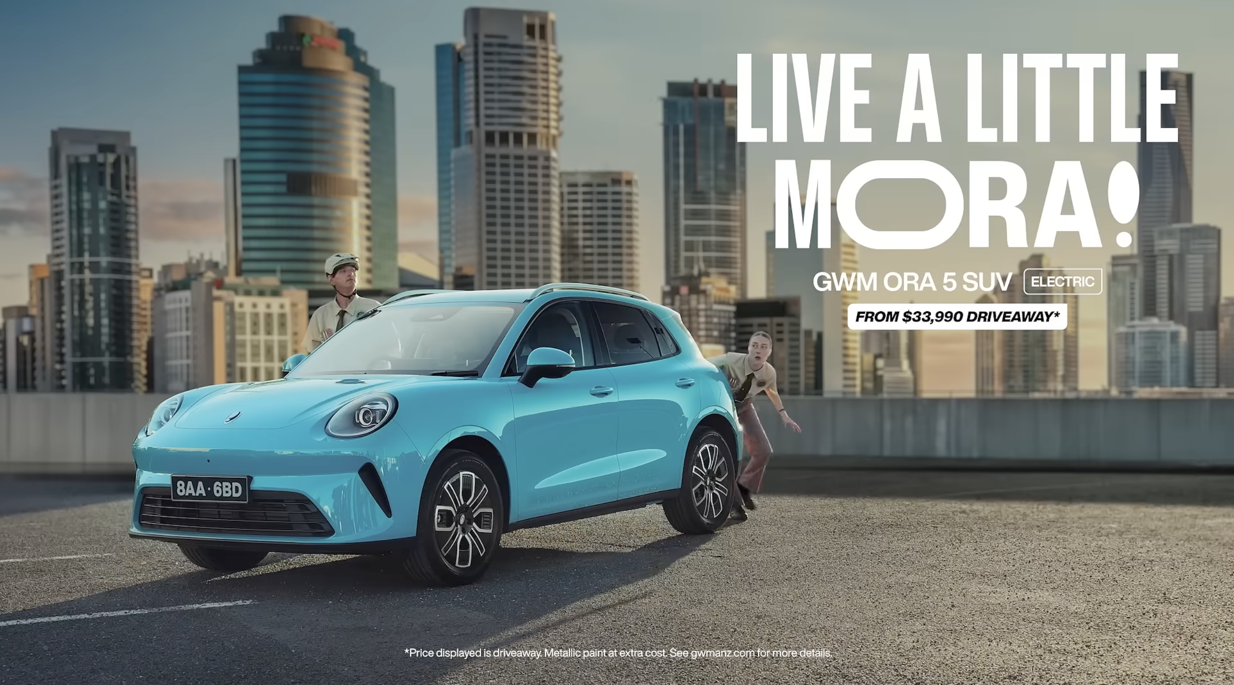 A bright blue electric SUV with a license plate 8AA 6BD parked on a city rooftop with a skyline of tall buildings in the background. Two children, one boy wearing a helmet and a girl, are interacting with the vehicle, and large text on the image promotes living a city life with this electric SUV, starting from $33,990.