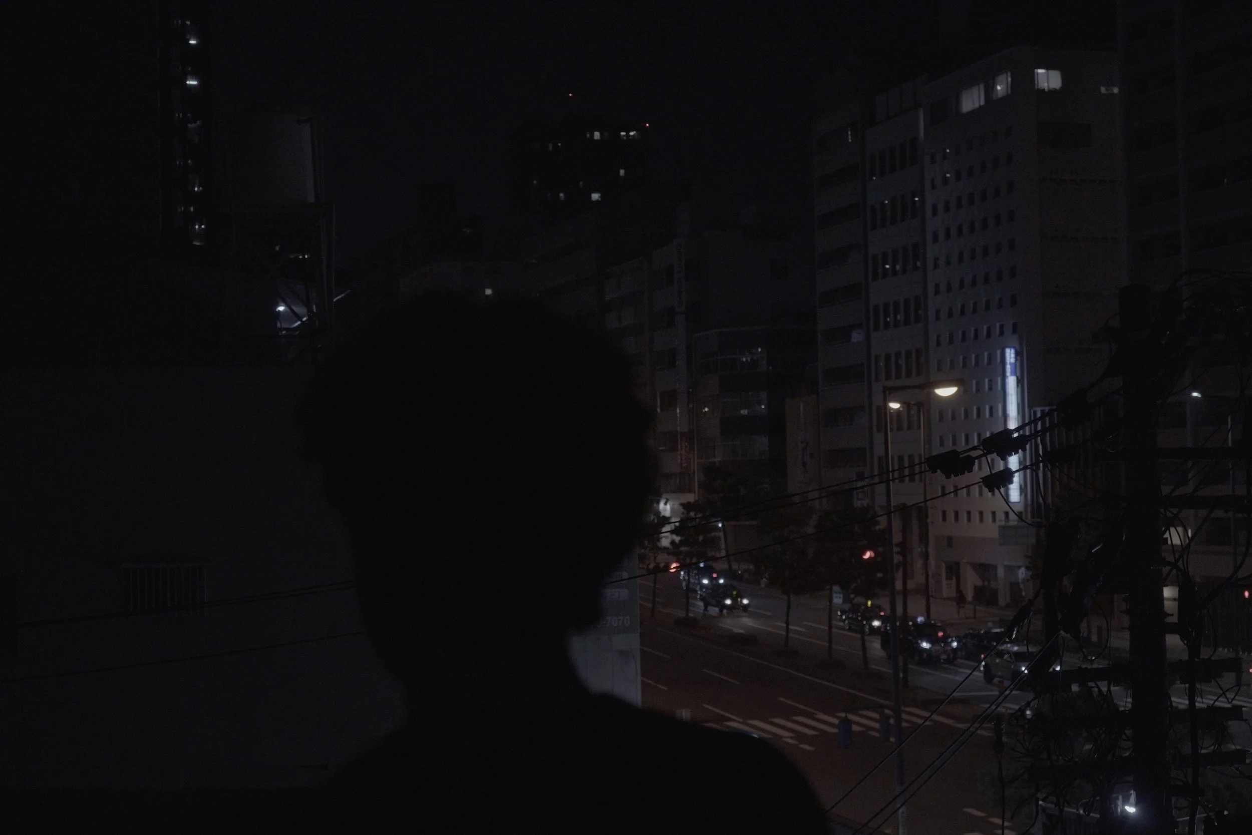 Night scene of city buildings with some lights on, a road with a few cars, and power lines in the foreground.