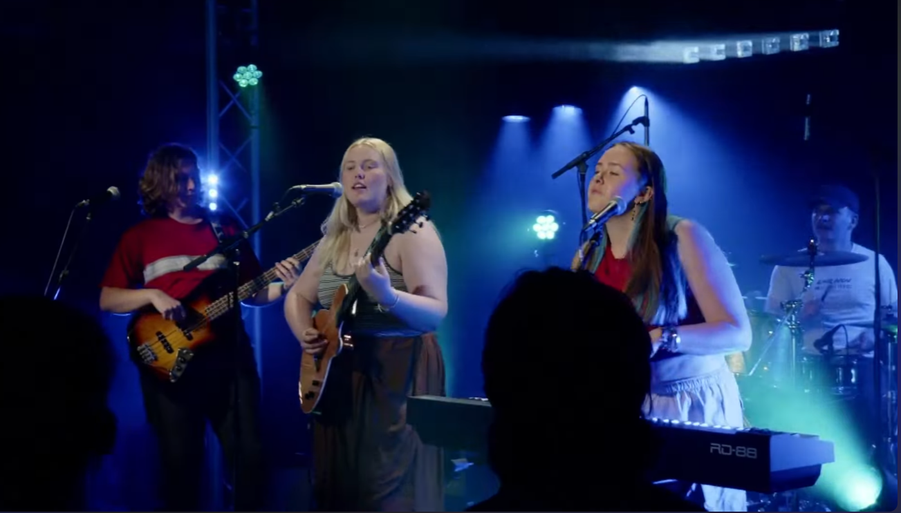Four young women performing on a stage with musical instruments, including guitars and drums, under colorful stage lights.