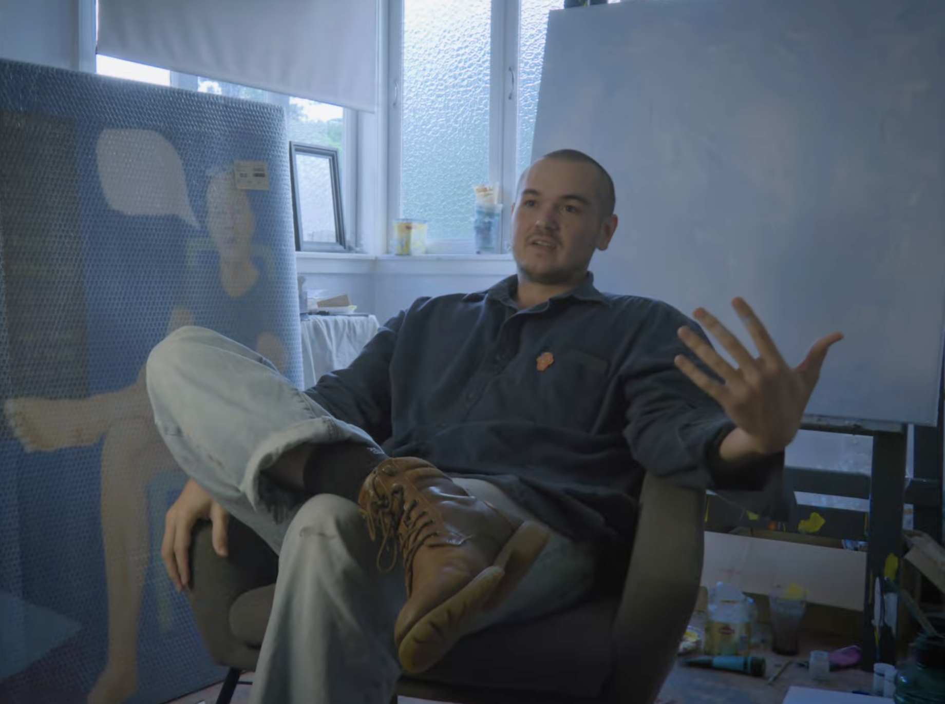 A man with a shaved head sitting in a chair, gesturing with his right hand while speaking in a brightly lit room with large windows, a window sill with items on it, and art supplies on the floor.